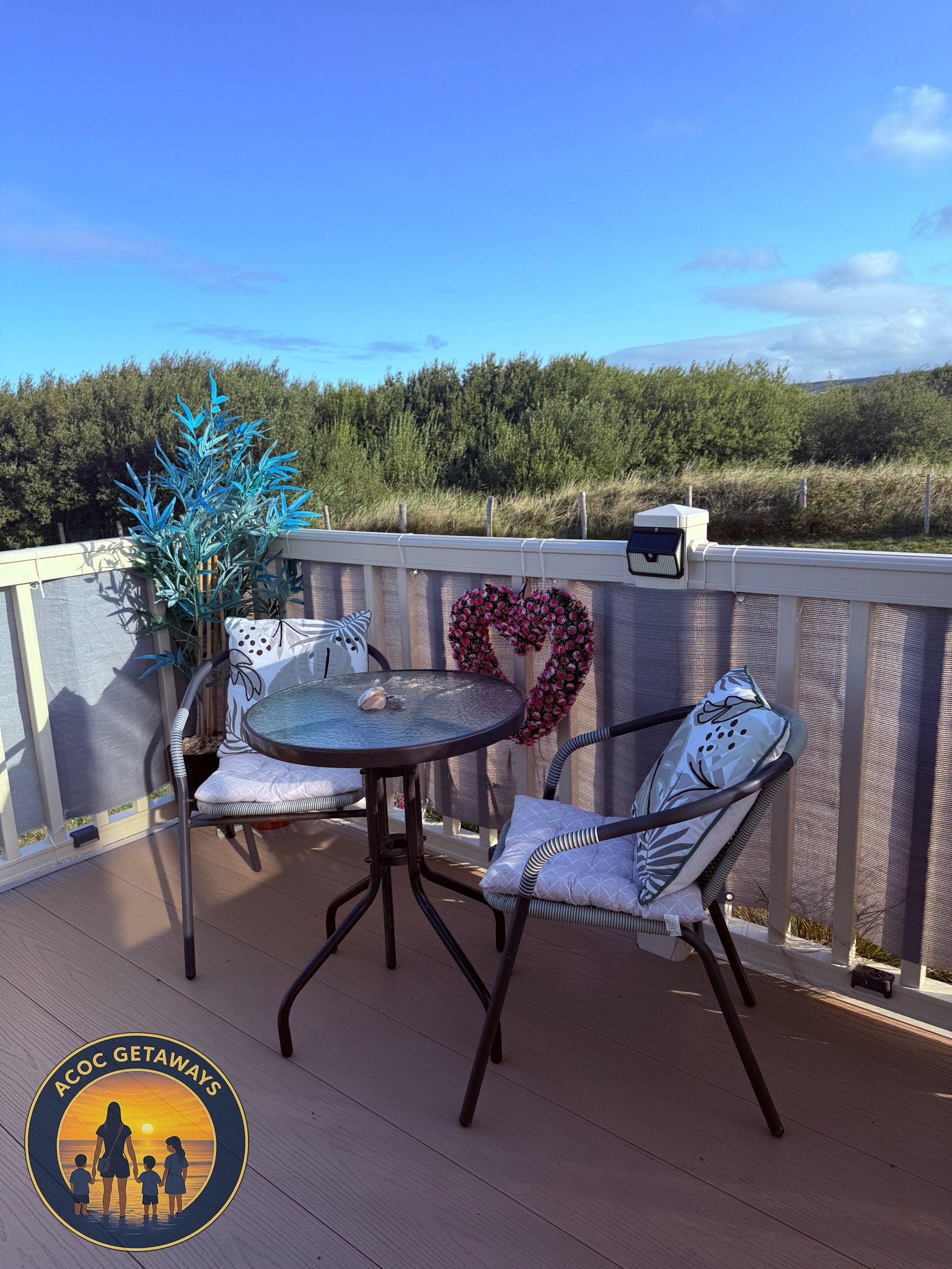 Outdoor balcony with two chairs, a glass table, decorative cushions, a blue potted plant, and a heart-shaped wreath made of pink flowers hanging on the railing, with a view of green trees and a blue sky.