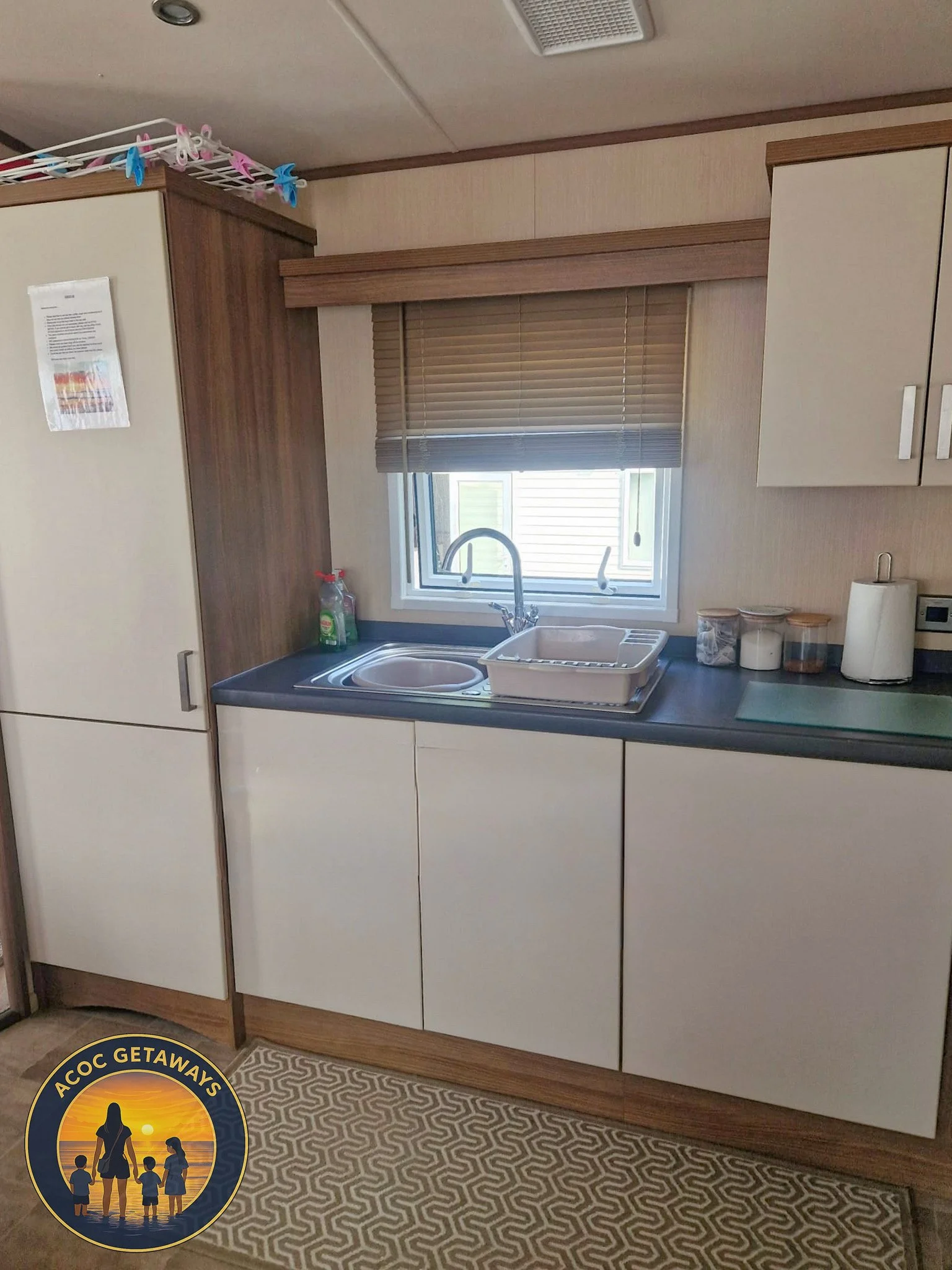 A small kitchen area with a window, sink, white cabinets, and a counter. There are some containers and a paper towel on the counter. A drying rack with dishes is above the sink, and a spray bottle is on the counter. The kitchen has a patterned rug on