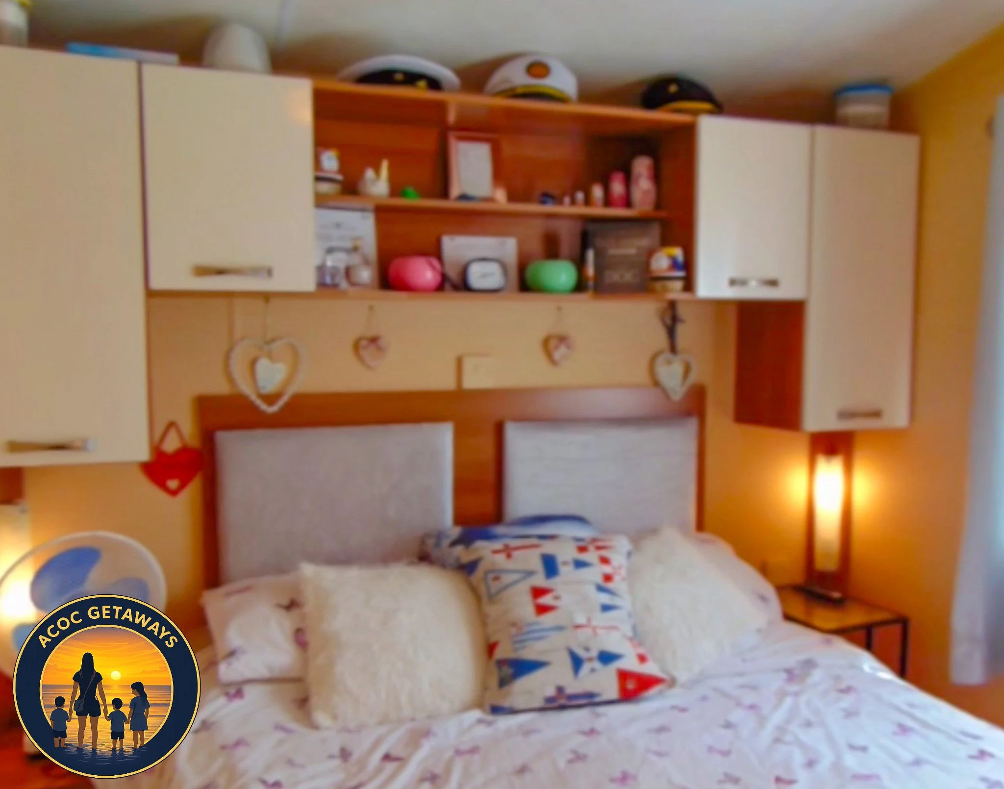 A cozy bedroom with a bed featuring a patterned quilt and decorative pillows, built-in wooden shelves behind the headboard displaying hats and various items, and a side table with a lamp on the right side of the bed. The wall is painted yellow, and h