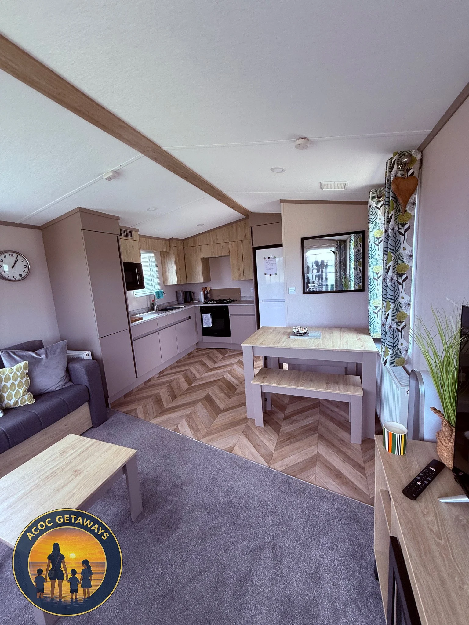 Living room with a gray sofa and wooden coffee table, open kitchen with cabinets, refrigerator, stove, and microwave, dining table with bench, window with patterned curtains, wall clock, and TV on a stand with remote and plant.