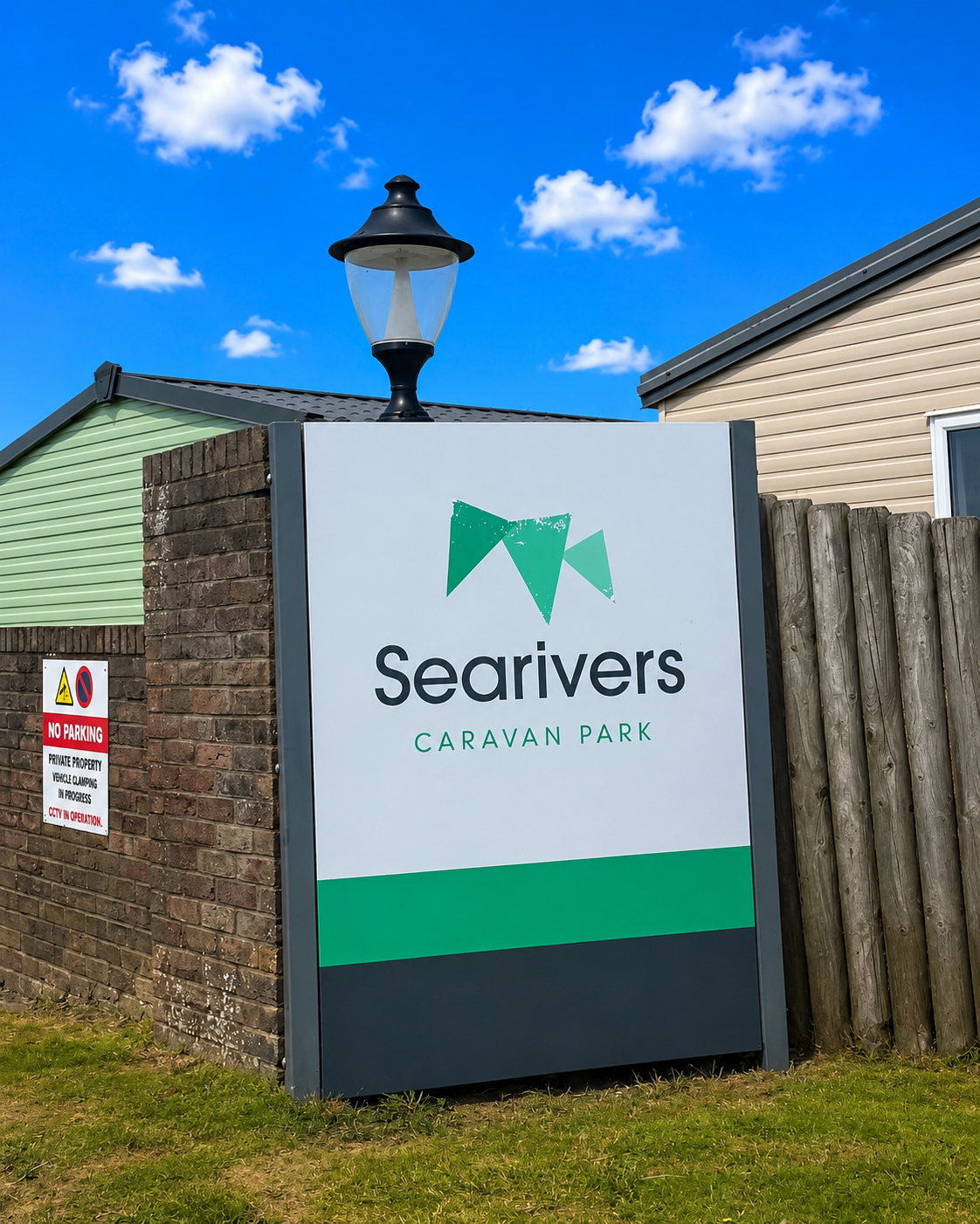 Sign for Searivers Caravan Park in front of brick and wood fencing, with a lamp post on top, under a blue sky with clouds.