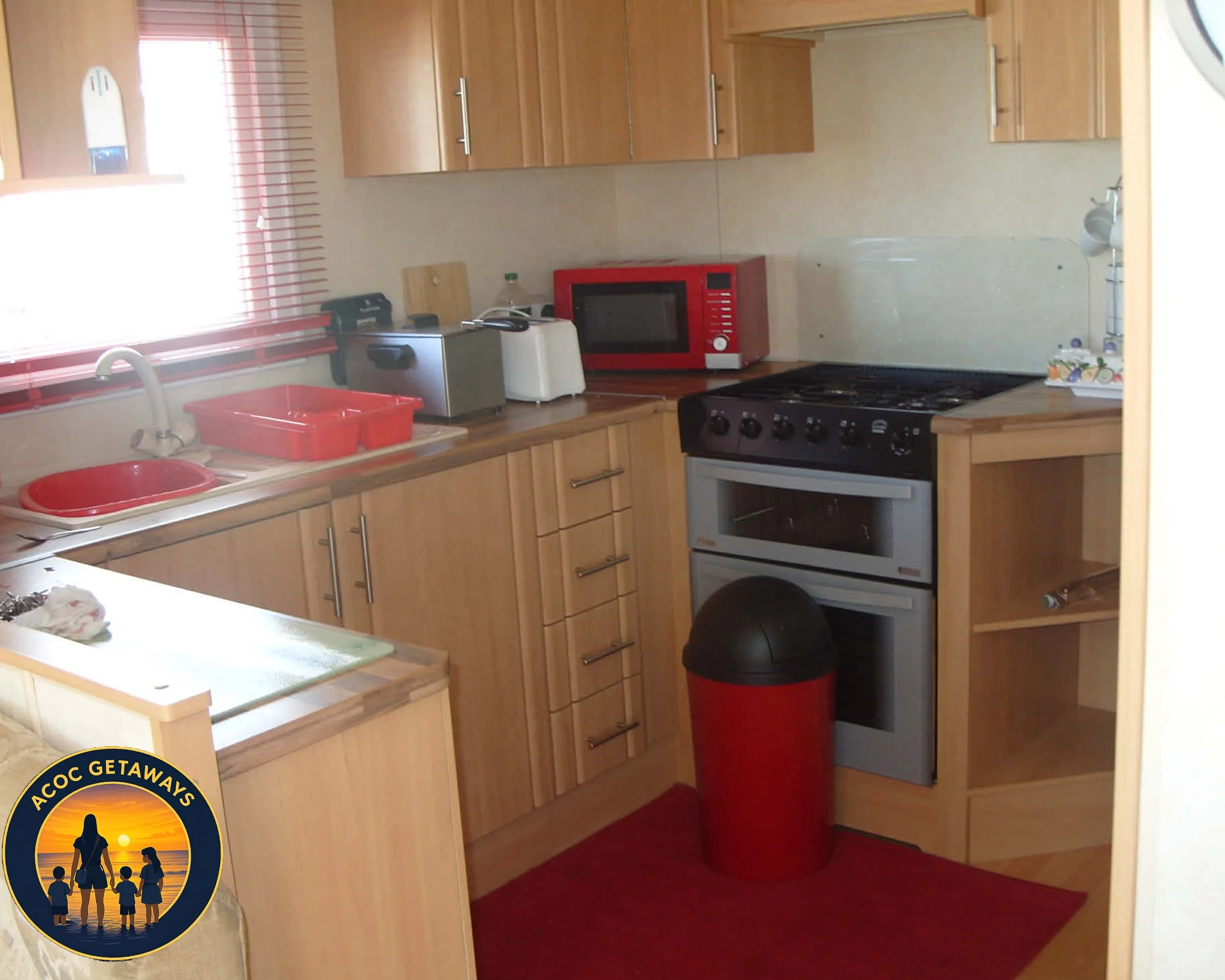 A compact kitchen with wooden cabinets, a red microwave, and a red trash bin. There are various kitchen appliances on the counter and a small window with red blinds.