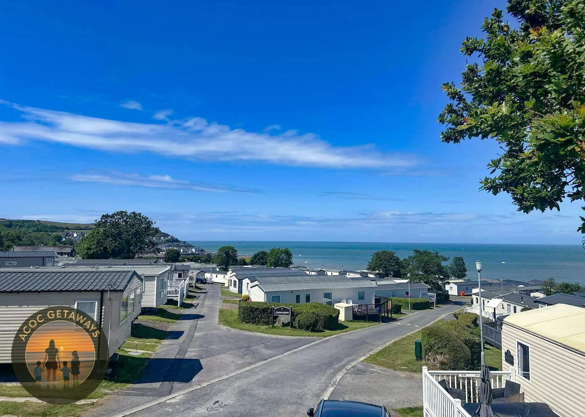 Beach view with mobile homes, trees, and ocean in the background under partly cloudy blue sky.