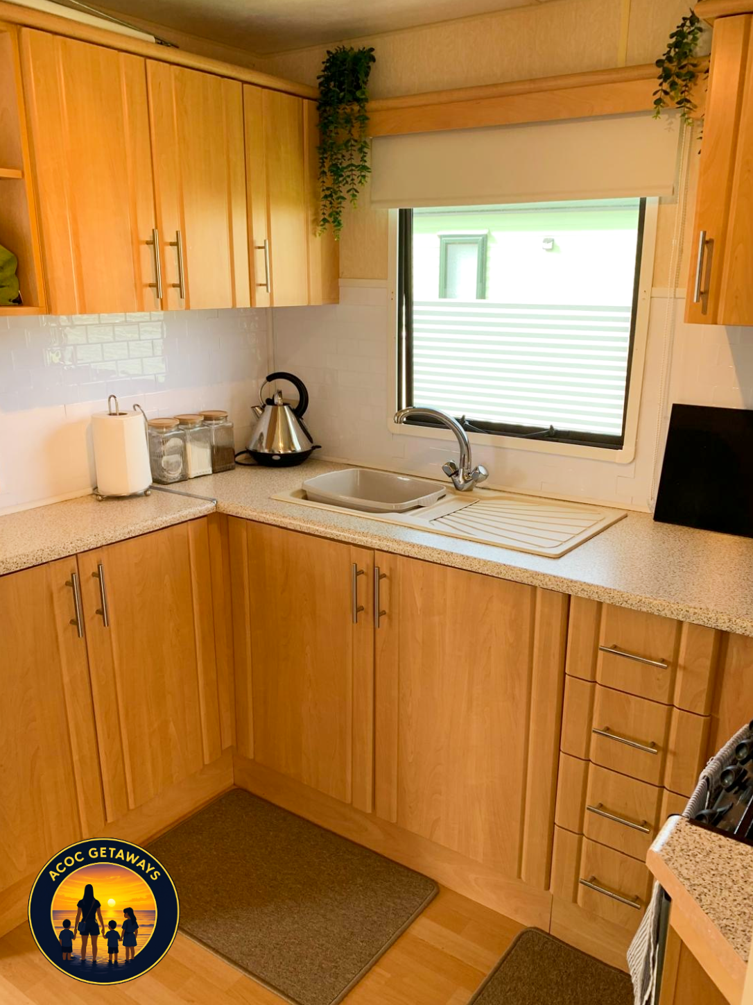 A small kitchen with wooden cabinets, beige countertops, a window with a view outside, a white sink with a silver faucet, a black and silver kettle, and kitchen canisters, with a brown mat on the floor.