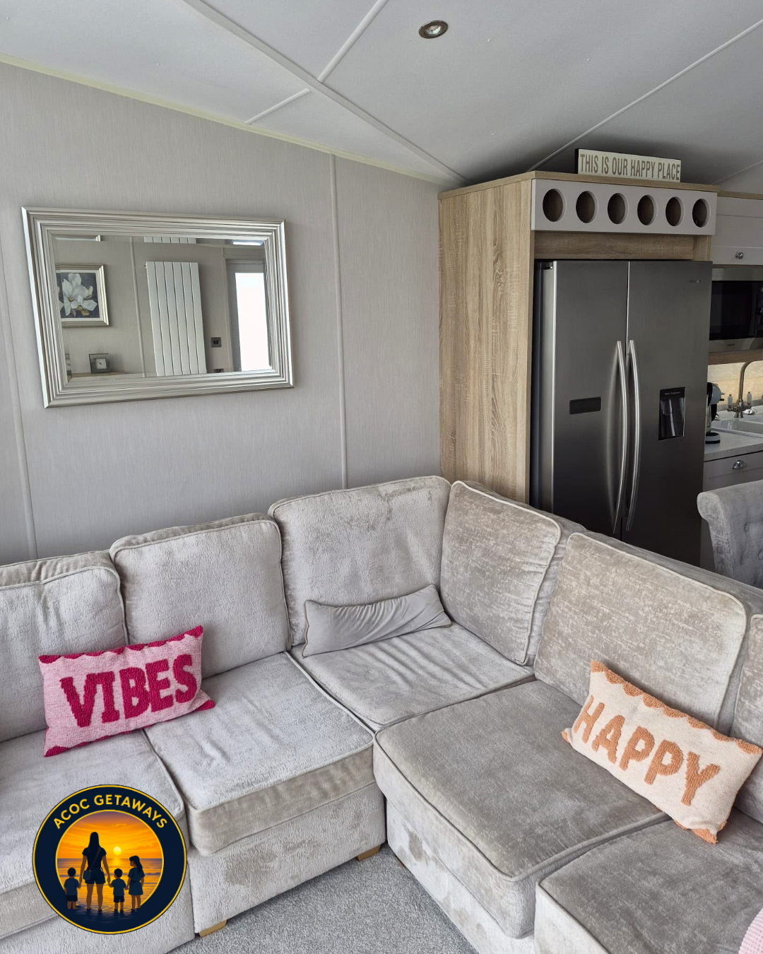Living room with beige sectional sofa, decorative pillows reading "VIBES" and "HAPPY", wall mirror, and wooden kitchen cabinet with a stainless steel fridge on the right.