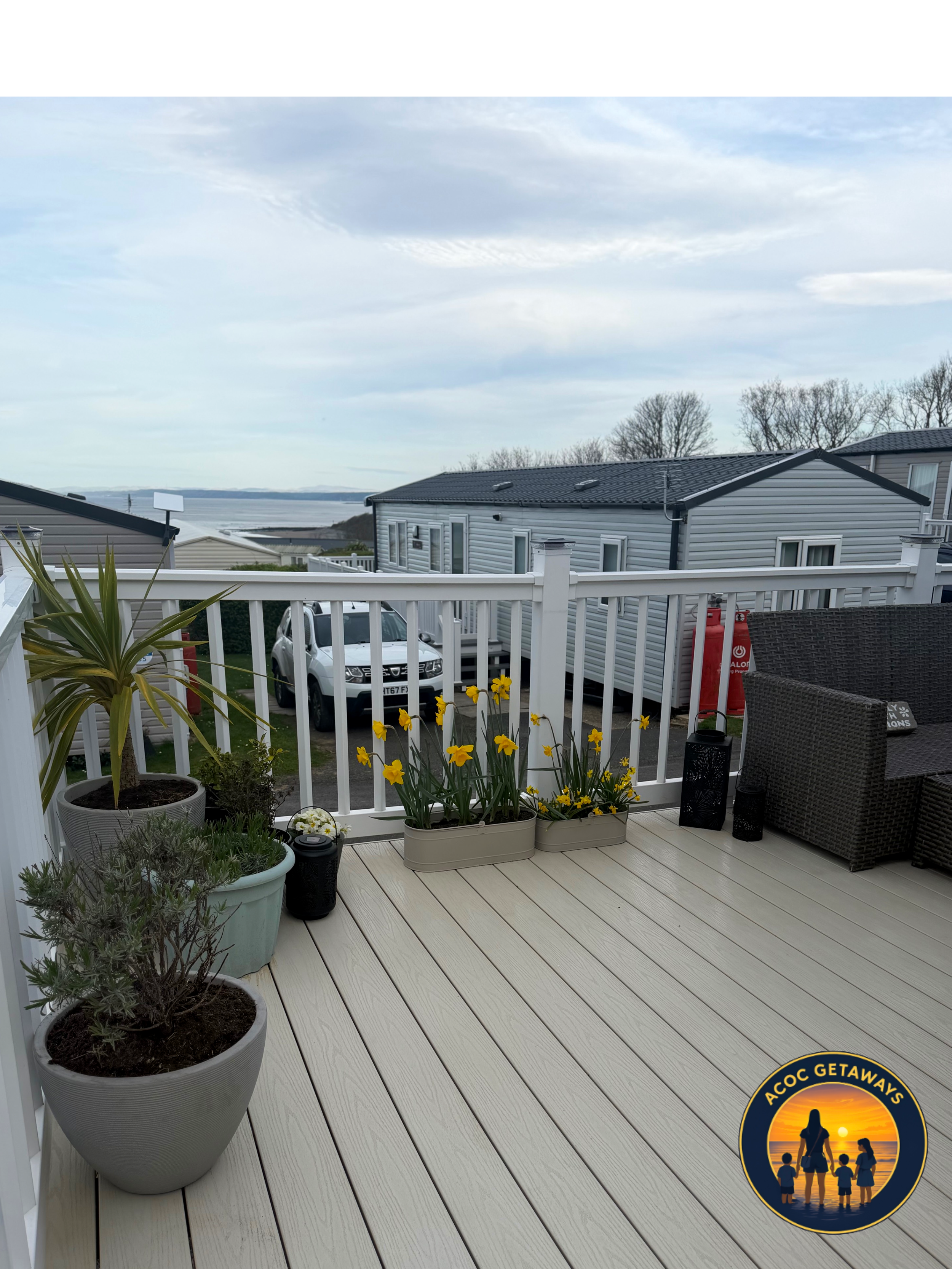 View of a white balcony with potted plants and flowers, overlooking houses and water in the distance under a cloudy sky.