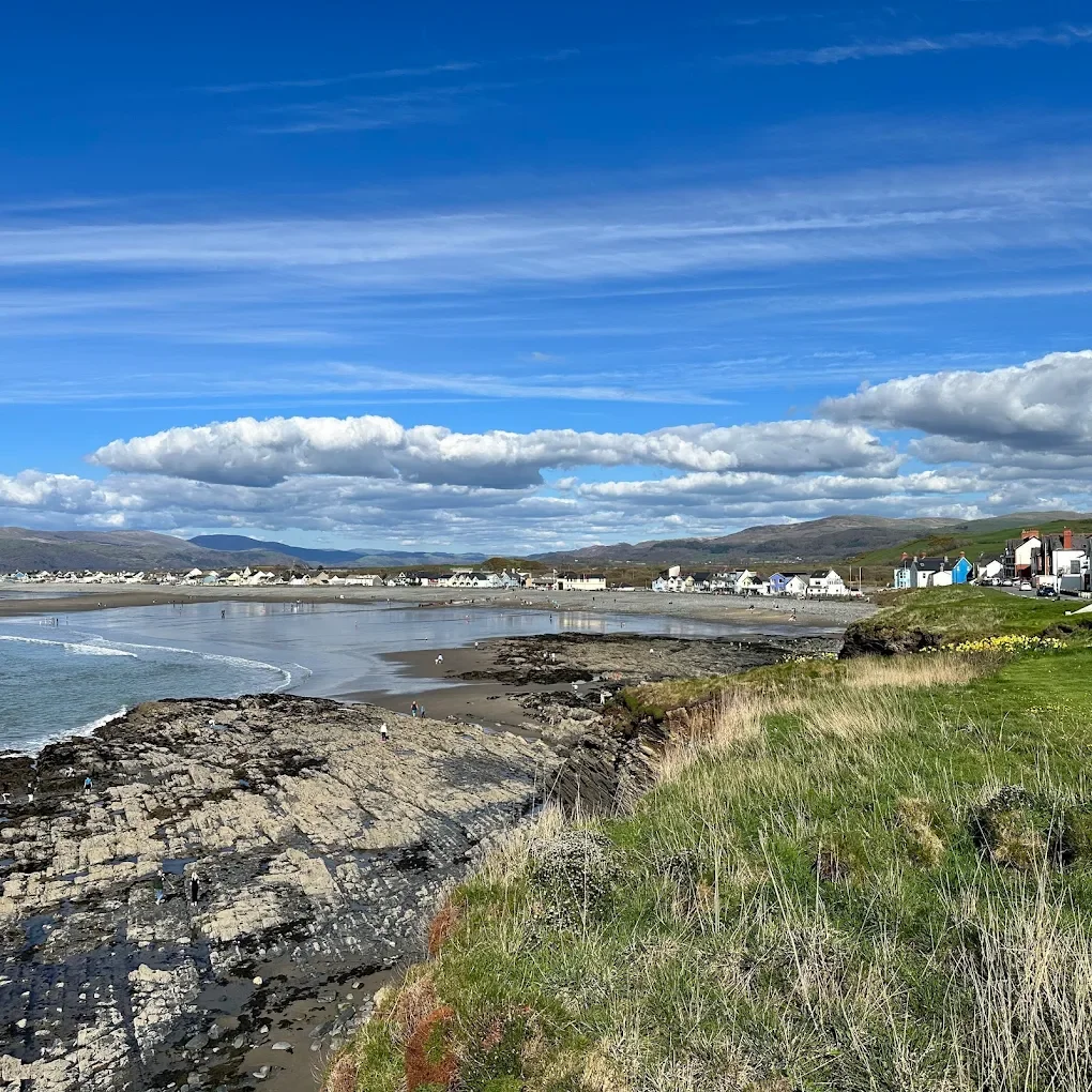 A coastal scene featuring a sandy beach with a rocky foreground, a small seaside town with colorful houses, and rolling hills in the background under a partly cloudy sky.