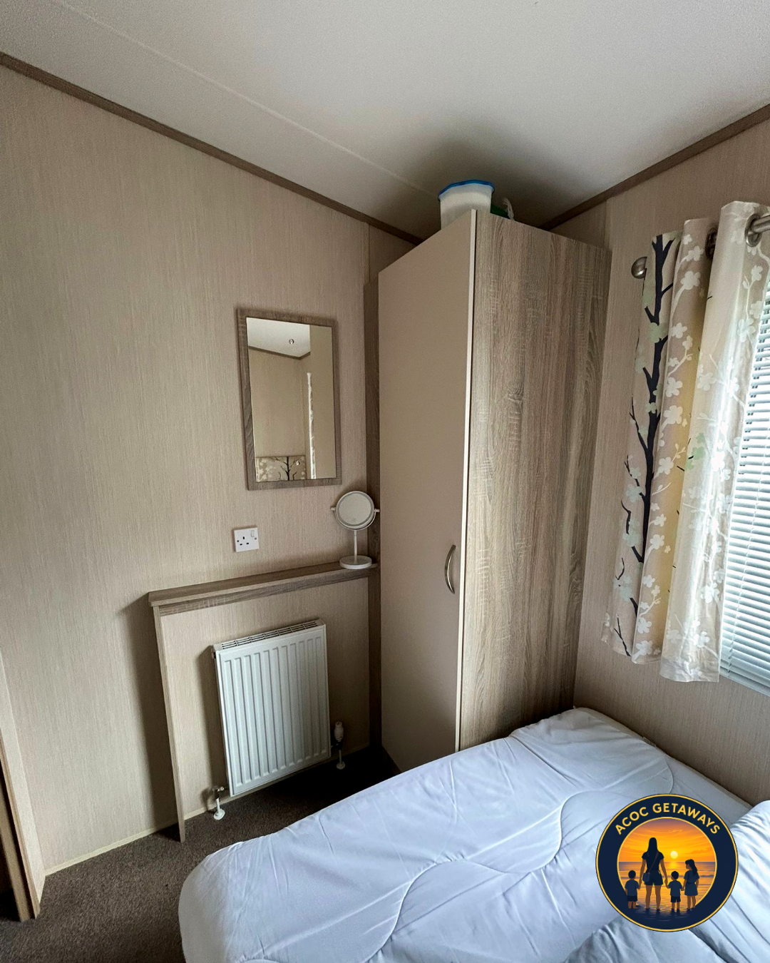 A bedroom with a bed, wardrobe, mirror, small round mirror, radiator, window with floral curtains, and a patterned wall.