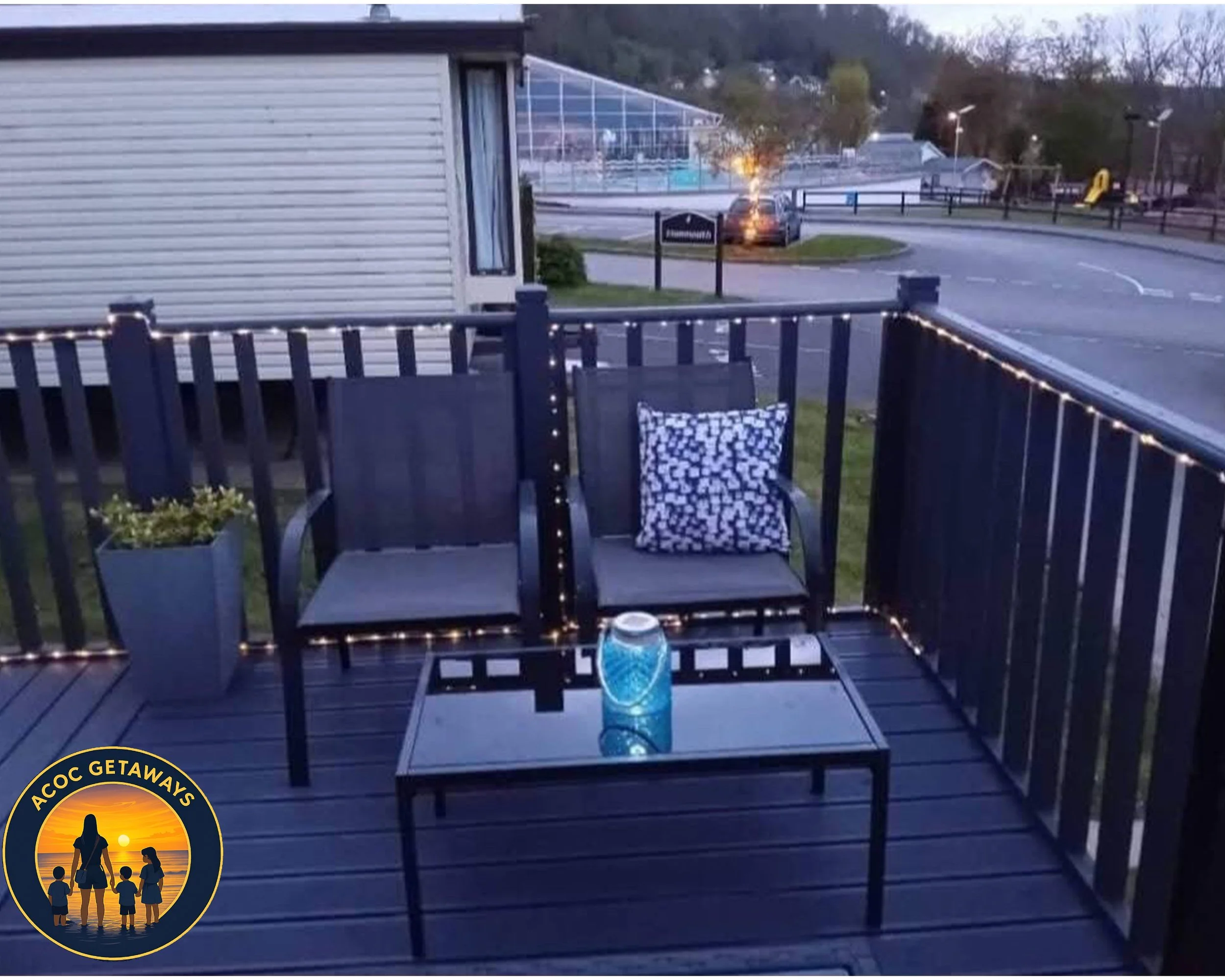 A small outdoor patio with two black chairs, one with a black and white patterned pillow, a small glass table with a blue lantern, and string lights along the black railing. In the background, a parking lot and tennis courts can be seen.