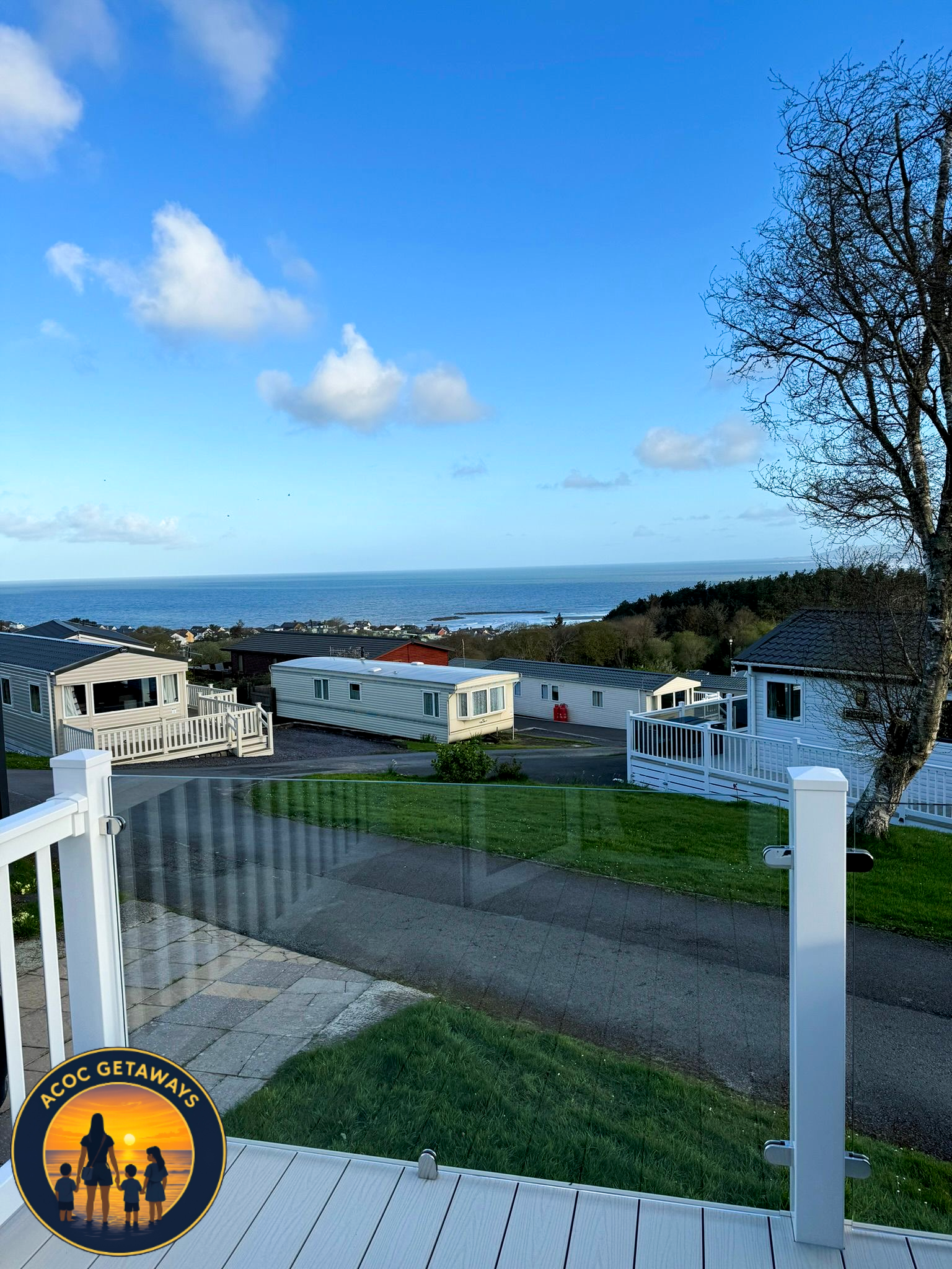 View of a coastal neighborhood with mobile homes, a grassy yard, a deck with a glass railing, a road, and the ocean in the distance under a blue sky with clouds.