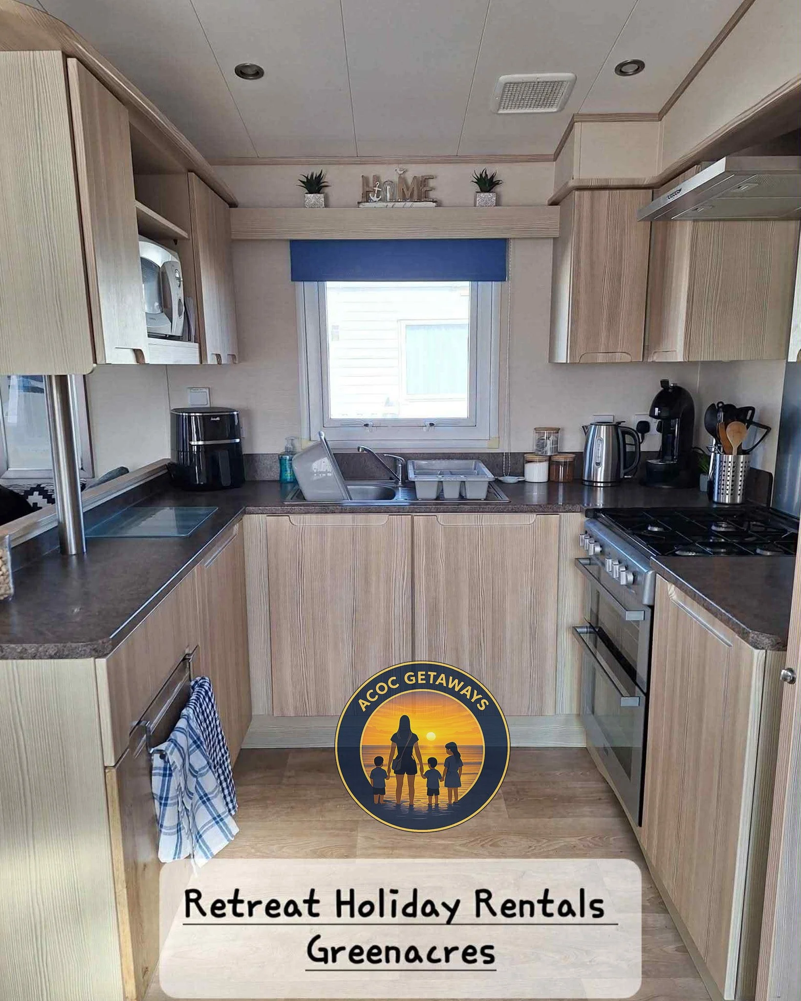 A kitchen with light wood cabinets, a window above the sink, and various appliances including a coffee maker, kettle, and microwave. Decor includes small potted plants and a shelf with 'LOVE' and 'HOME' signs. There is also a circular logo with a sil