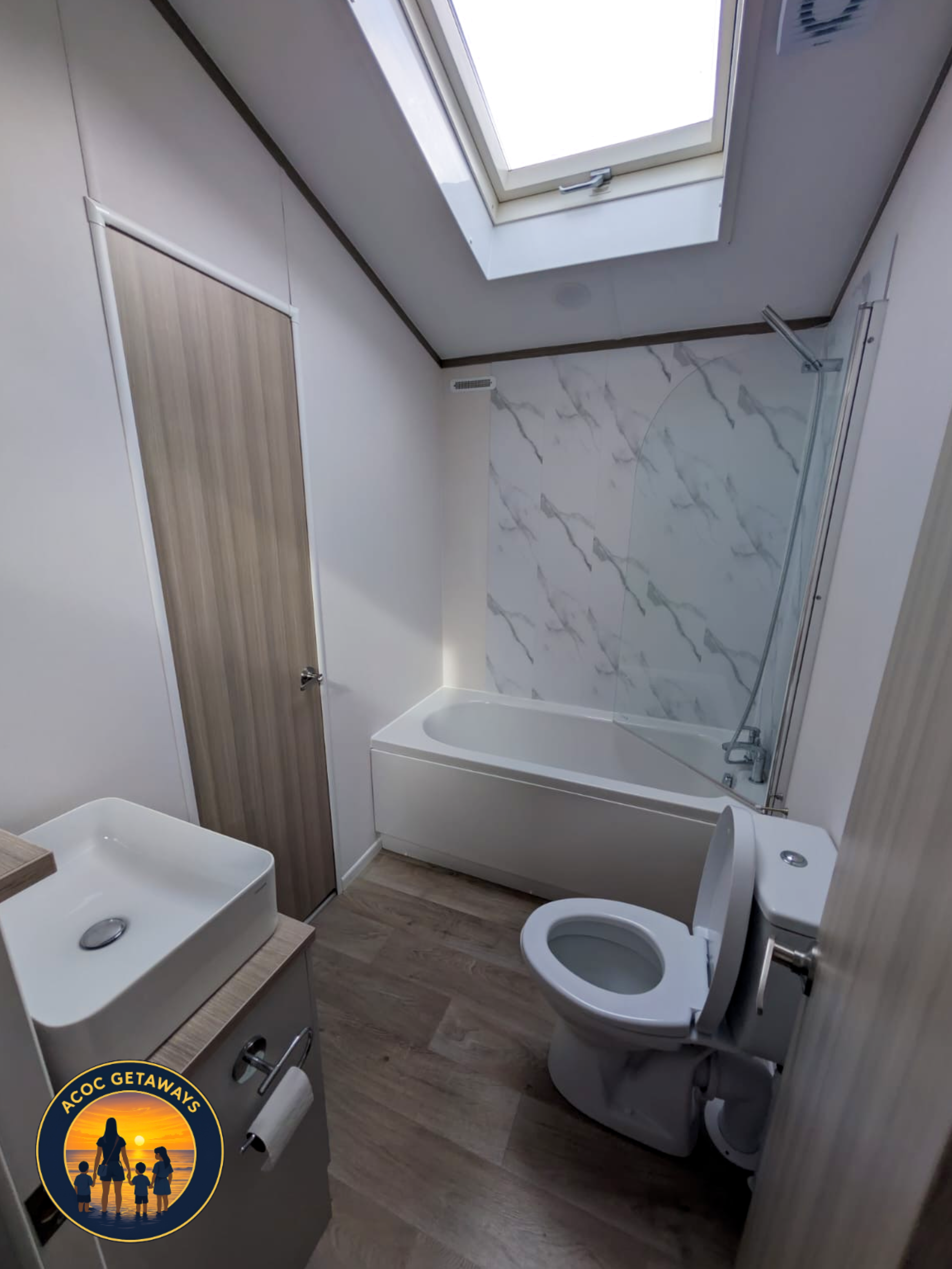 Small bathroom with a skylight window, a bathtub with a white marble tile wall, a toilet, and a small sink with a cabinet underneath. There is a wooden door and wood-style flooring.