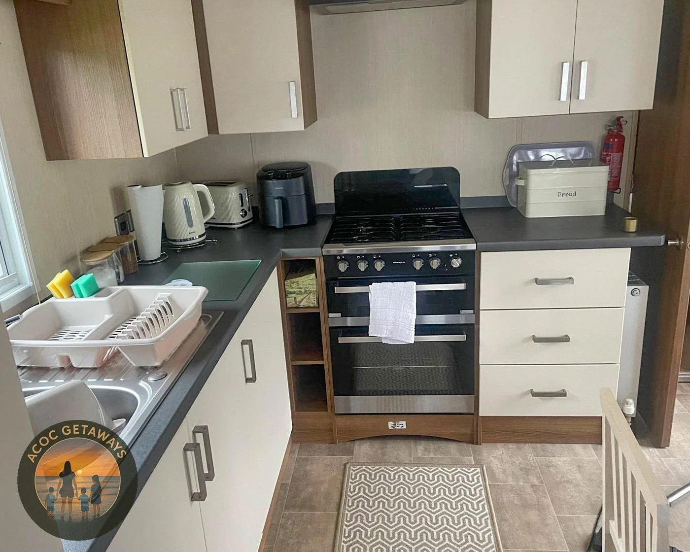 Kitchen with white cabinets, black countertops, and a stove in the center. Items include a dish drying rack with dish soap, a sink, several small appliances like a toaster, kettle, and air fryer, and a bread box. There is a window on the left and a fire extinguisher on the right.