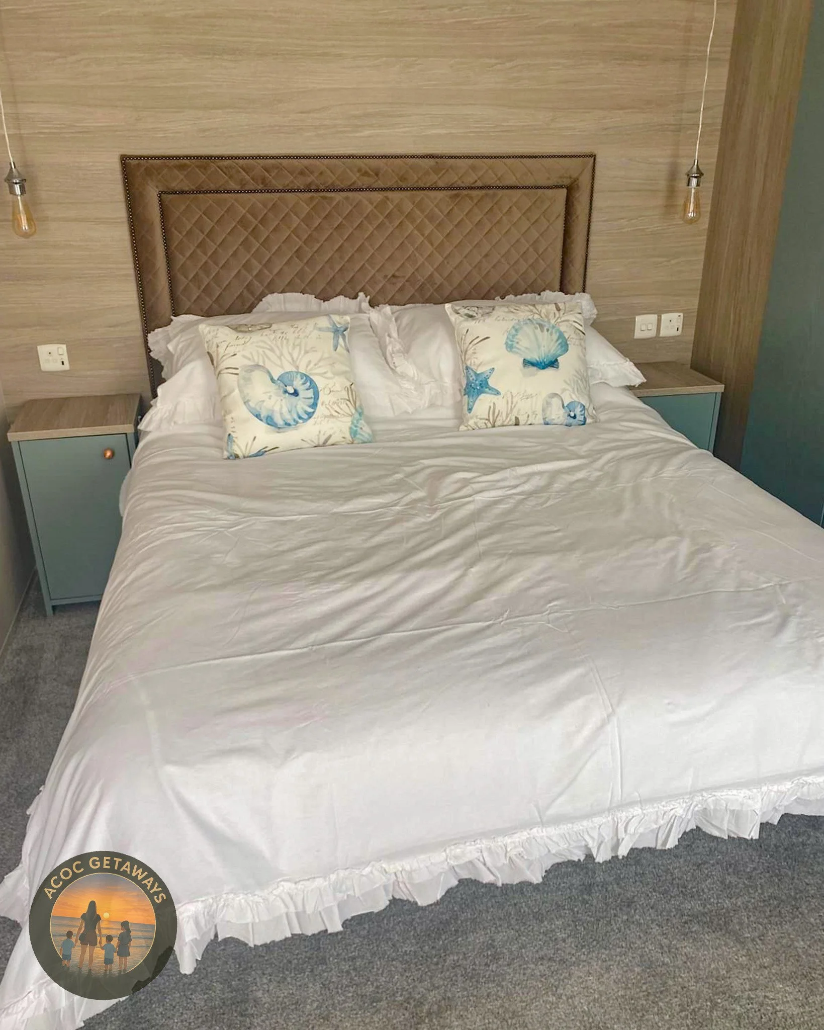 A neatly made bed with a padded beige headboard, white ruffled bed skirt, and four pillows featuring beach and shell designs in a hotel room with wooden walls and bedside tables on each side.