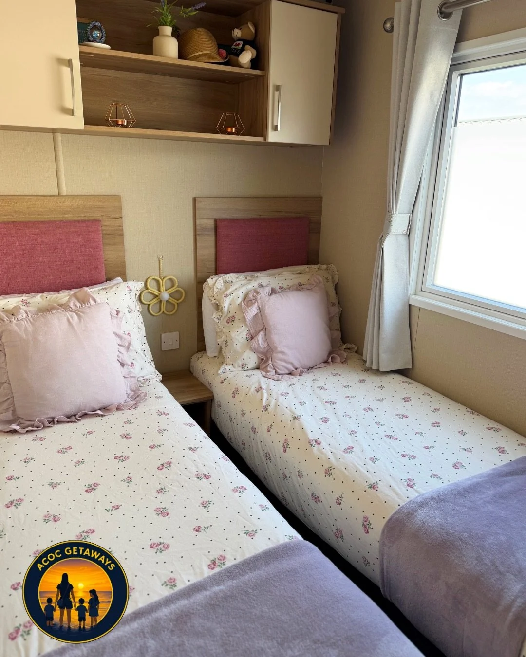 Two single beds with pink pillows and floral bedding in a bedroom, a window with curtains, and a wooden shelf above the beds with decorative items.