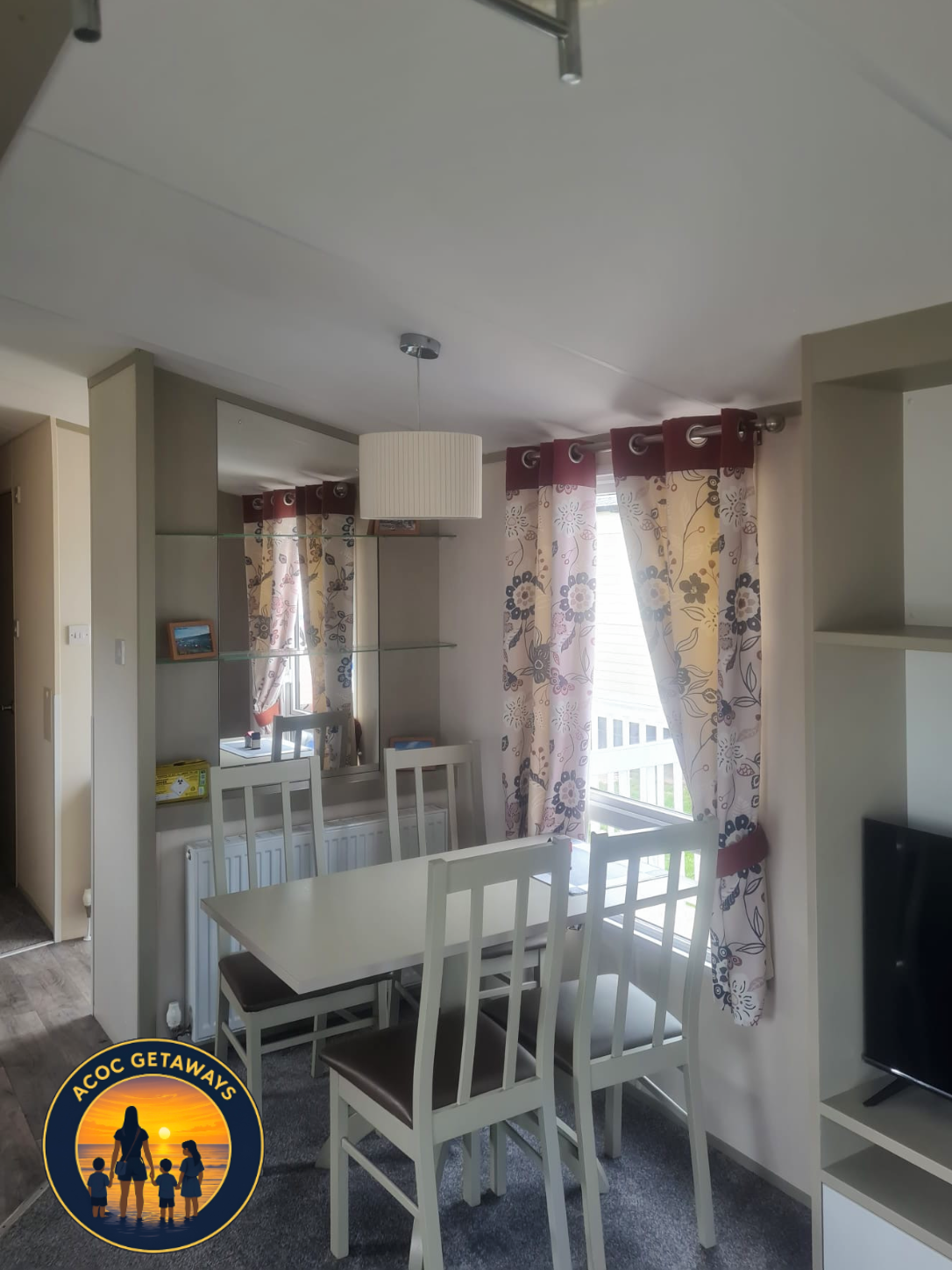 Dining area with a white table and four white chairs, floral curtains covering windows, a ceiling pendant light, and a decorative mirror on the wall.