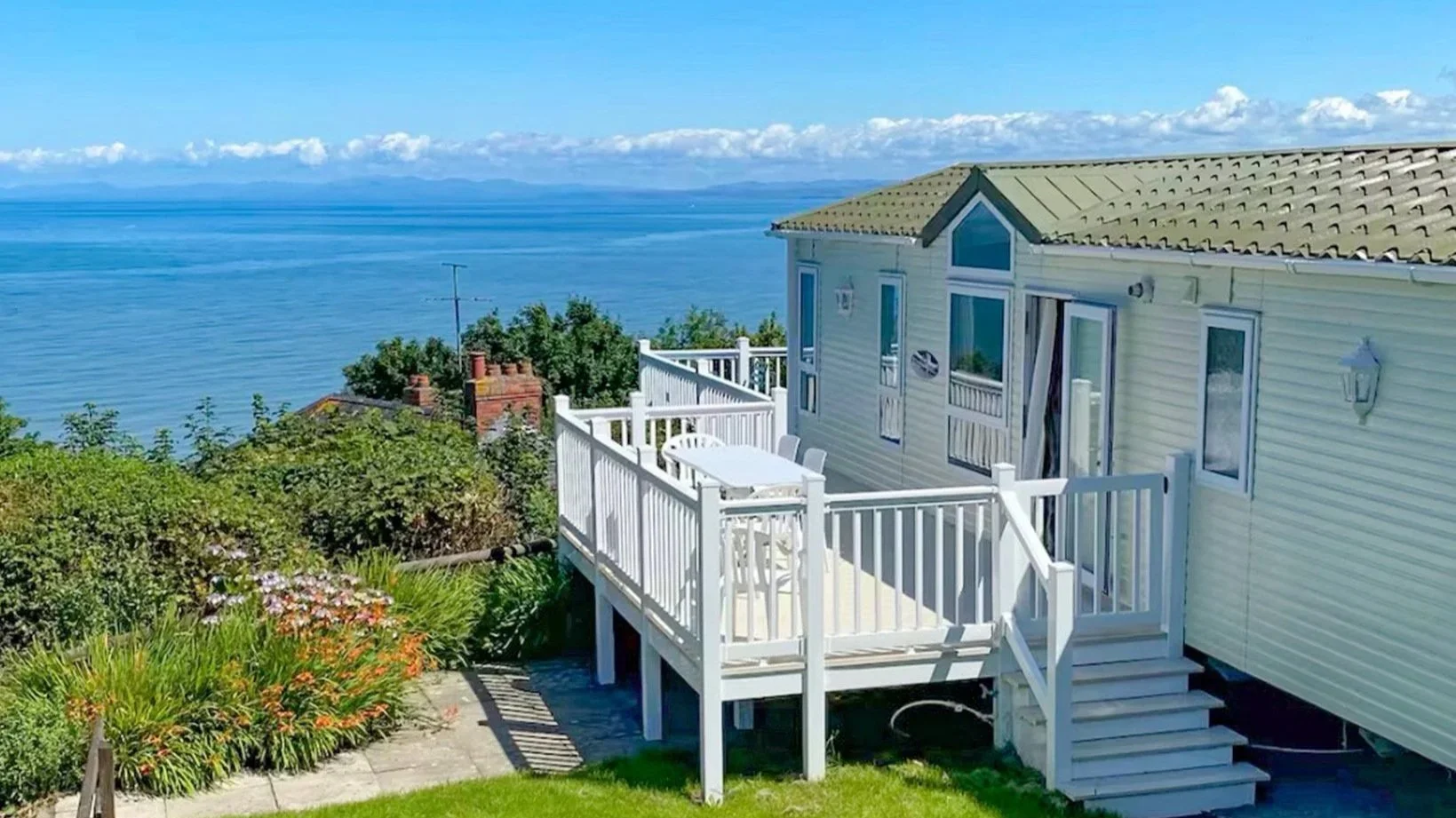 A house with a white deck overlooking a body of water and a distant mountain range in the background, with landscaped garden plants in the foreground.