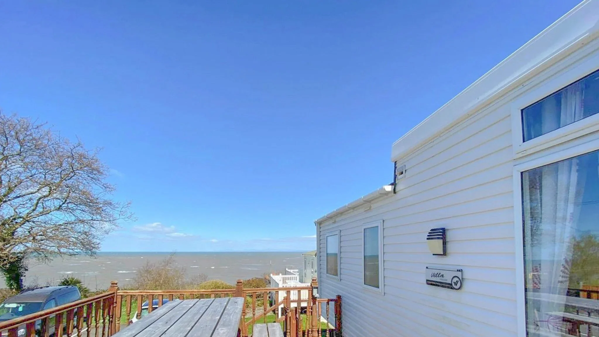 View of a house with beige siding, a deck with a wooden railing, and an ocean in the background under a clear blue sky.