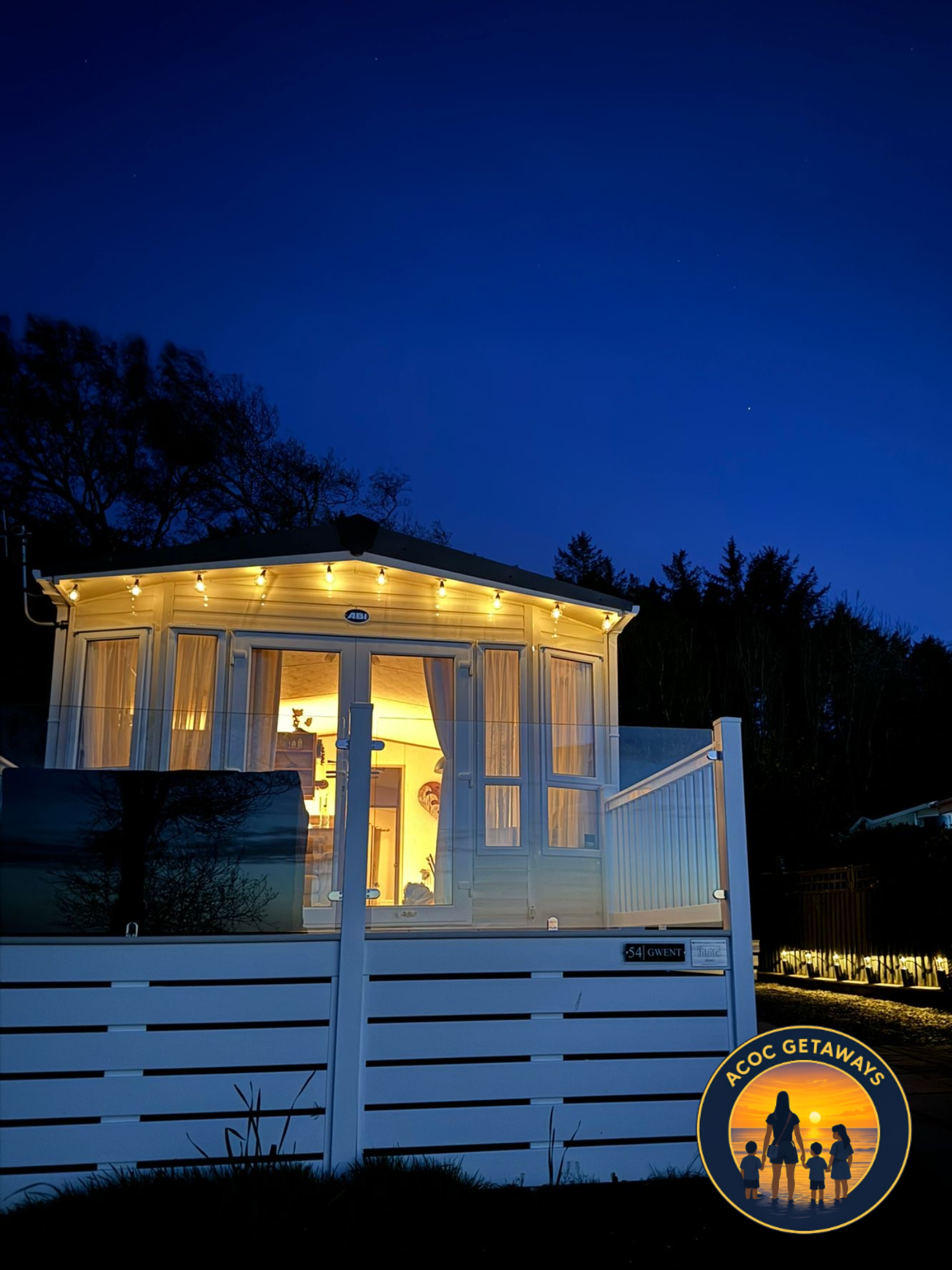 A cozy house with large windows and a porch, illuminated from inside, outside at night with a dark blue sky and trees in the background.