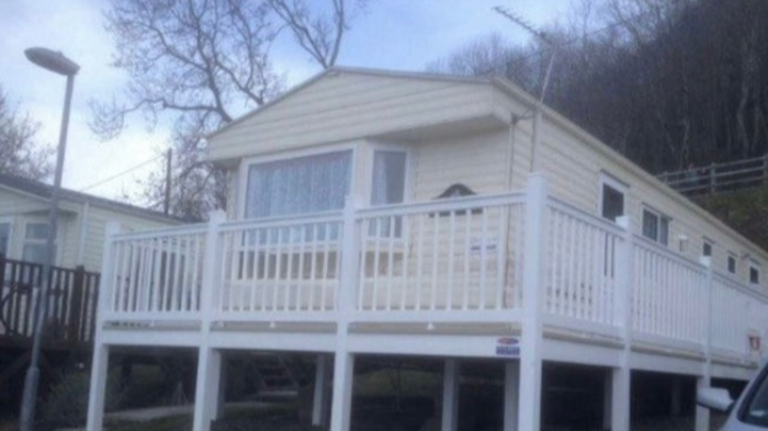 A house with beige siding, a white porch railing, and a large window, situated on a grassy hillside with bare trees in the background.