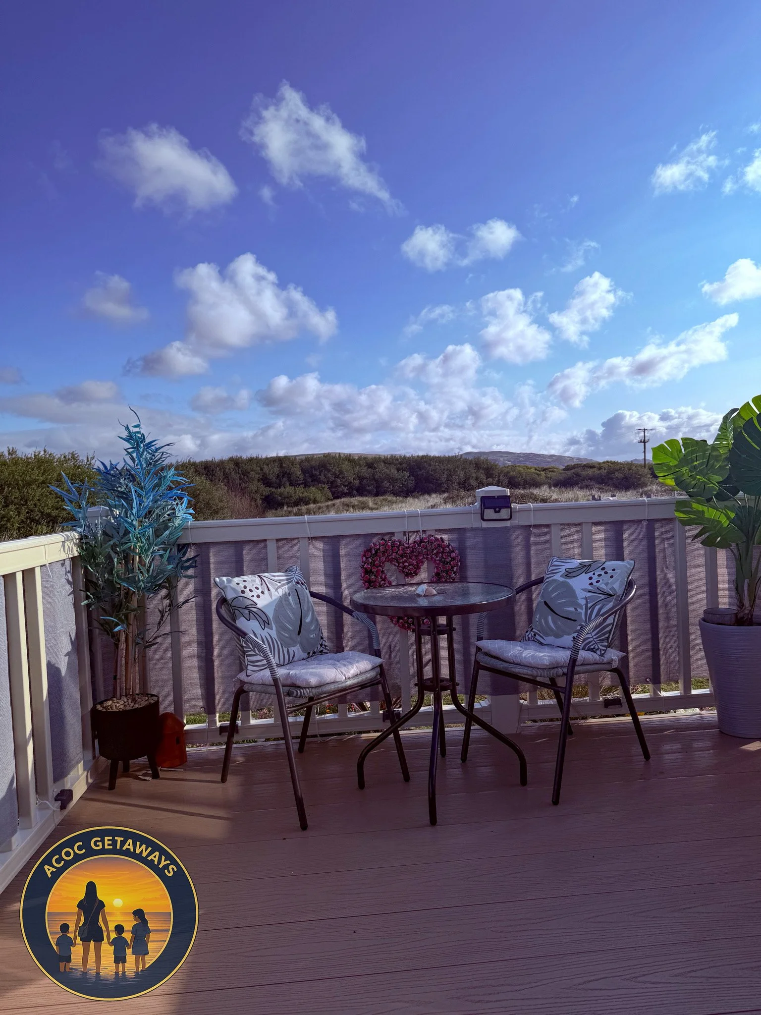 A wooden deck outdoors with two chairs that have floral cushions, a round table, green potted plants, a heart-shaped wreath, and a scenic view of trees and a blue sky with clouds in the background.