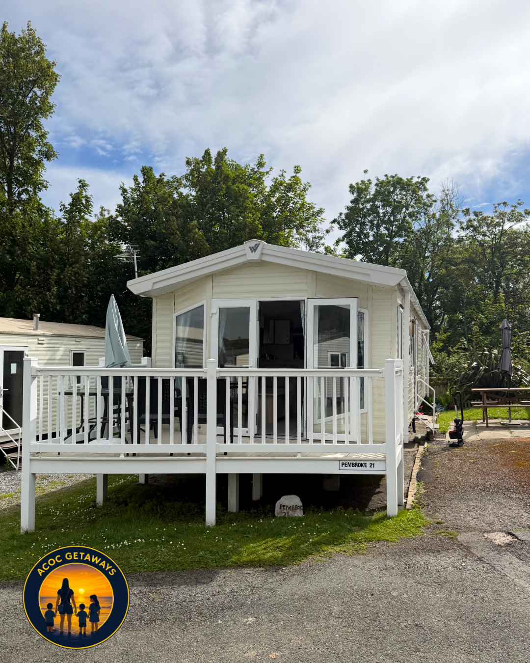 White mobile home with a front porch and pillars, surrounded by trees and a partly cloudy sky.