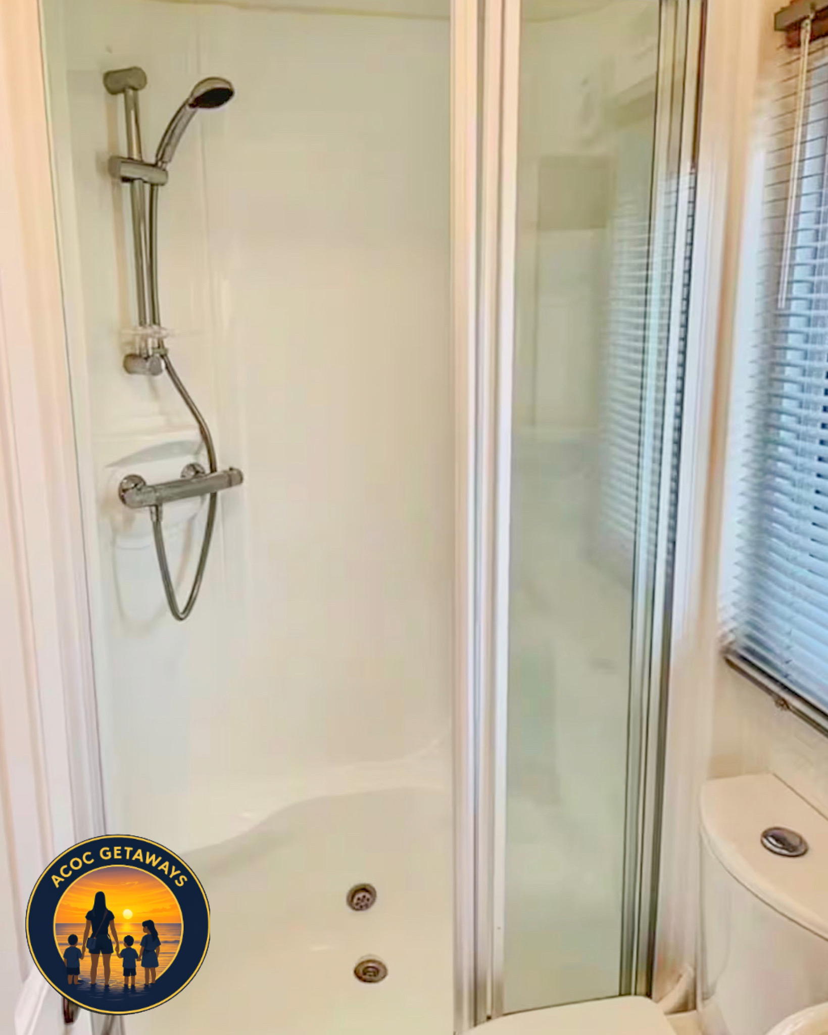 The image shows a closed glass shower door with a silver showerhead and fixtures inside. There is a window with blinds to the right of the shower. A logo with the text "ACOC GETAWAYS" and an illustration of a family at sunset is overlayed at the bott