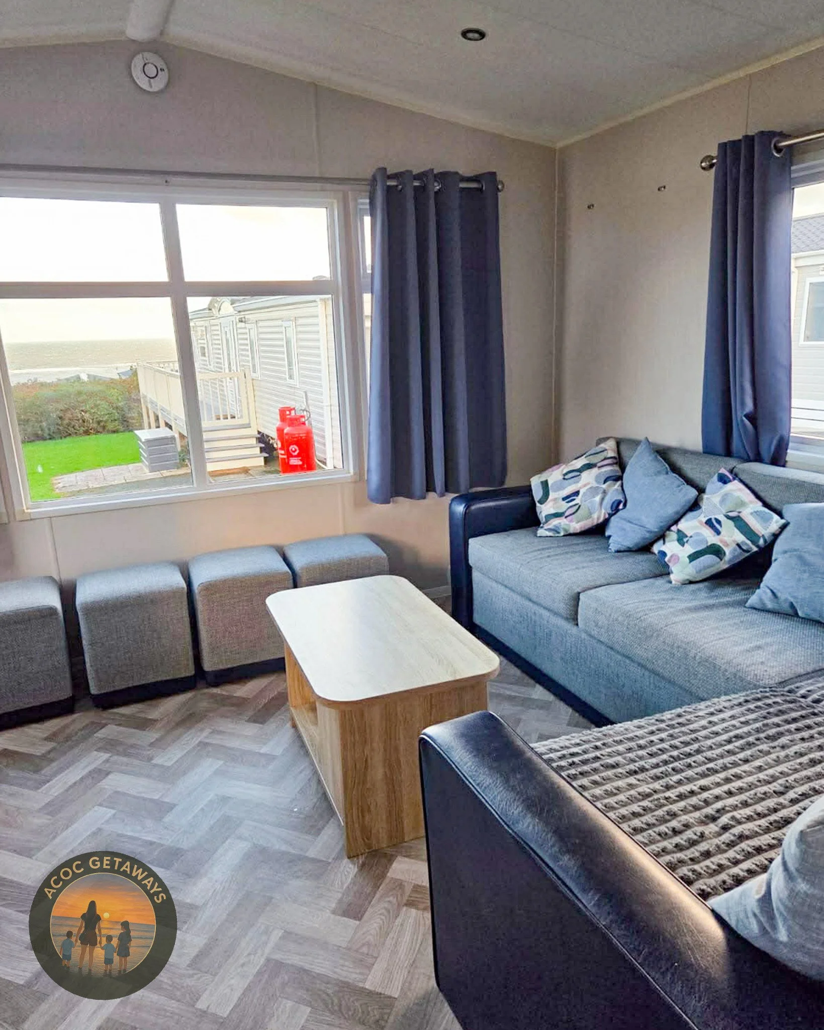 A cozy living room with gray sofa, several colorful pillows, blue curtains, a wooden coffee table, and a window showing a view of outdoor stairs, a fire extinguisher, and a grassy yard.