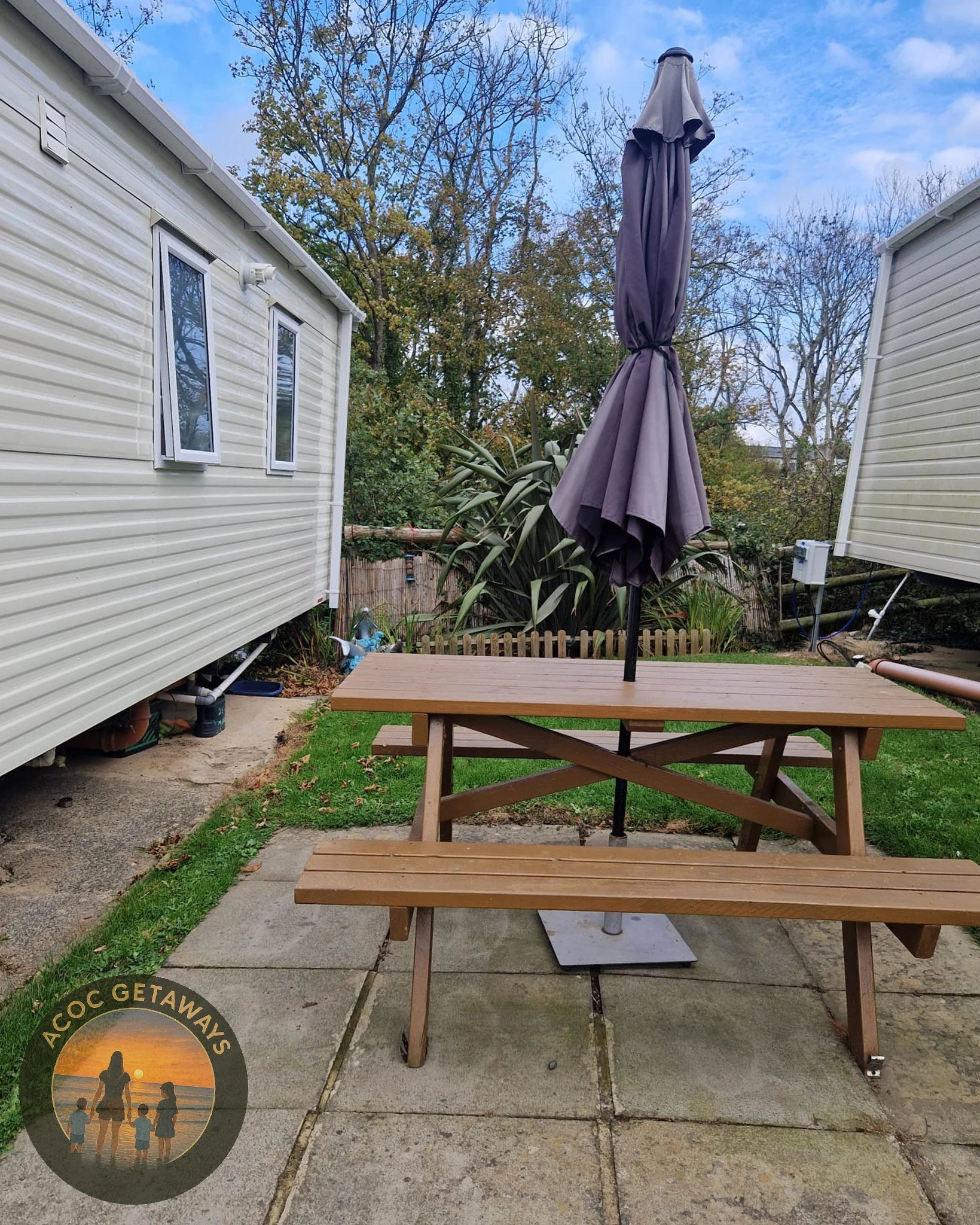 Wooden picnic table with attached bench on a concrete patio, topped with a closed purple outdoor umbrella. The background features a fence, trees, and a mobile home or trailer with white siding.