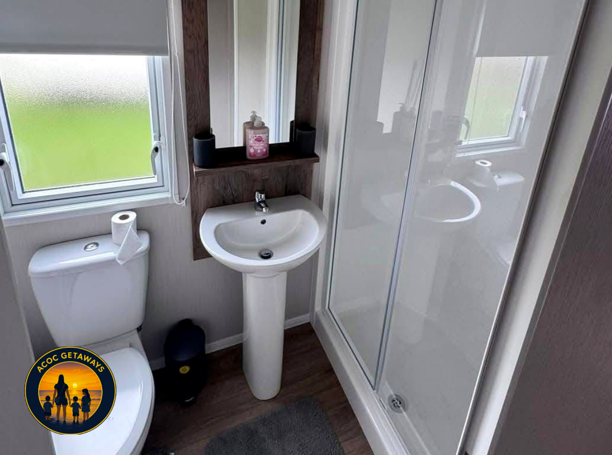 Small bathroom with a white toilet, a white pedestal sink, a mirror, a window with frosted glass, a soap dispenser, a small shelf, and a shower with a glass door.