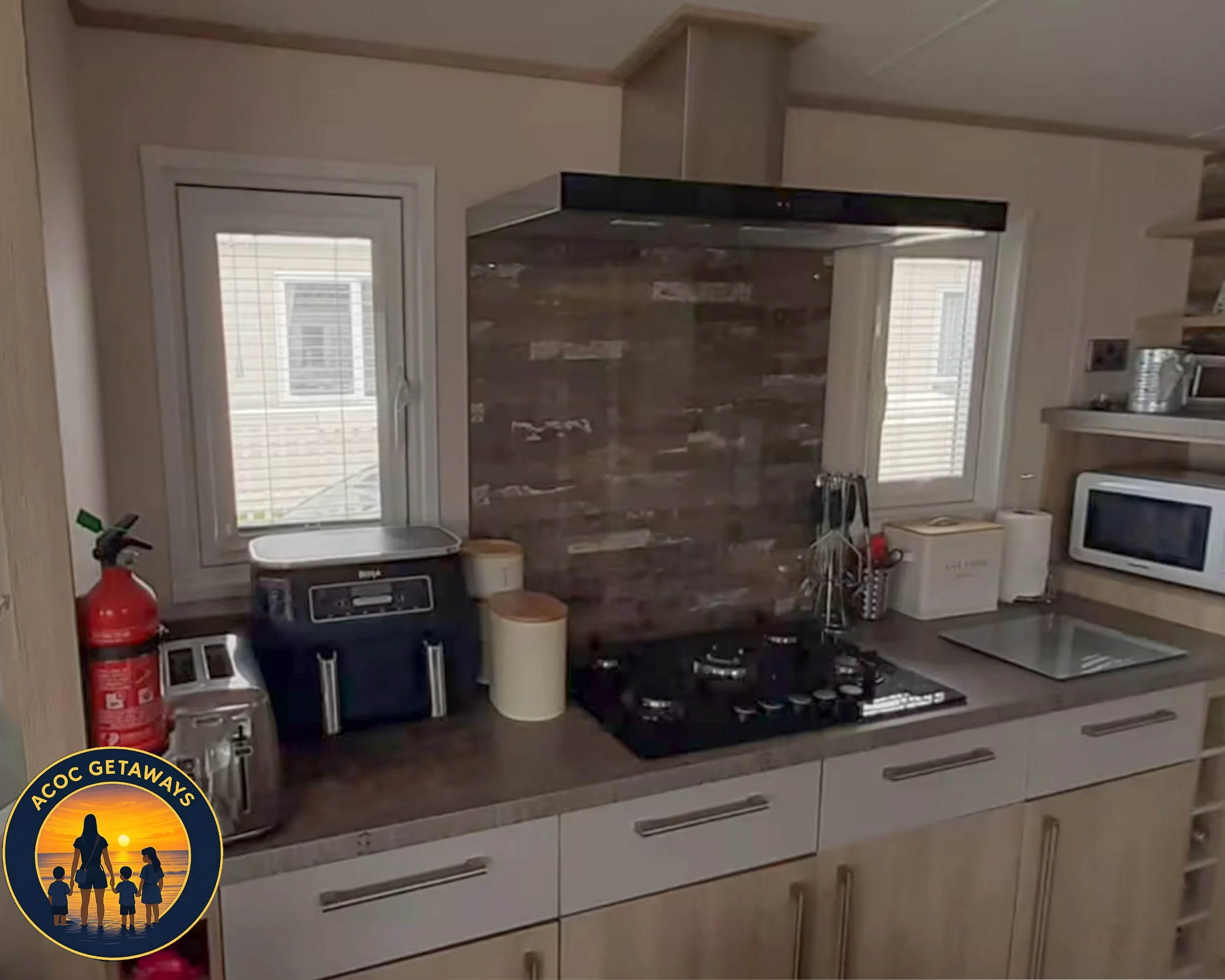 A kitchen countertop with a fire extinguisher, toaster, bread bins, coffee maker, and paper towel roll. There are two small windows above the countertop, a cooktop, microwave, and wooden shelves on the right side.