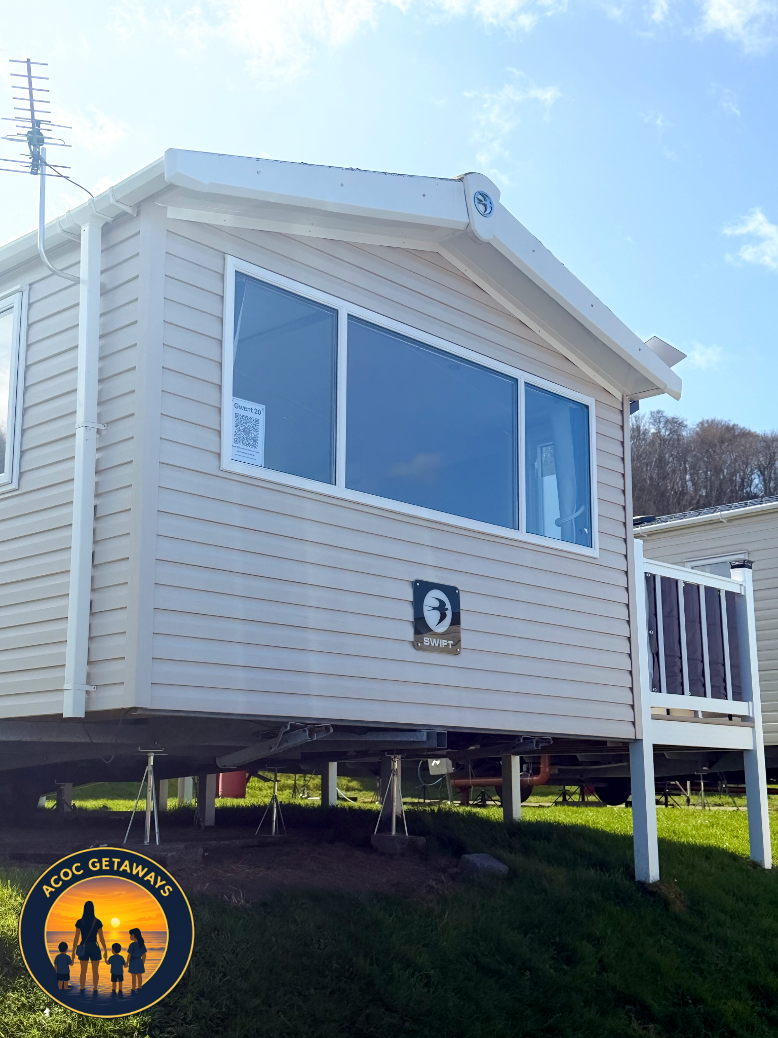A beige tiny house on stilts with large front window, a small balcony, and an A-frame roof. There is a small logo of a bird on a black background on the house, and a VIN number sticker on the window. An antenna is mounted on the roof."