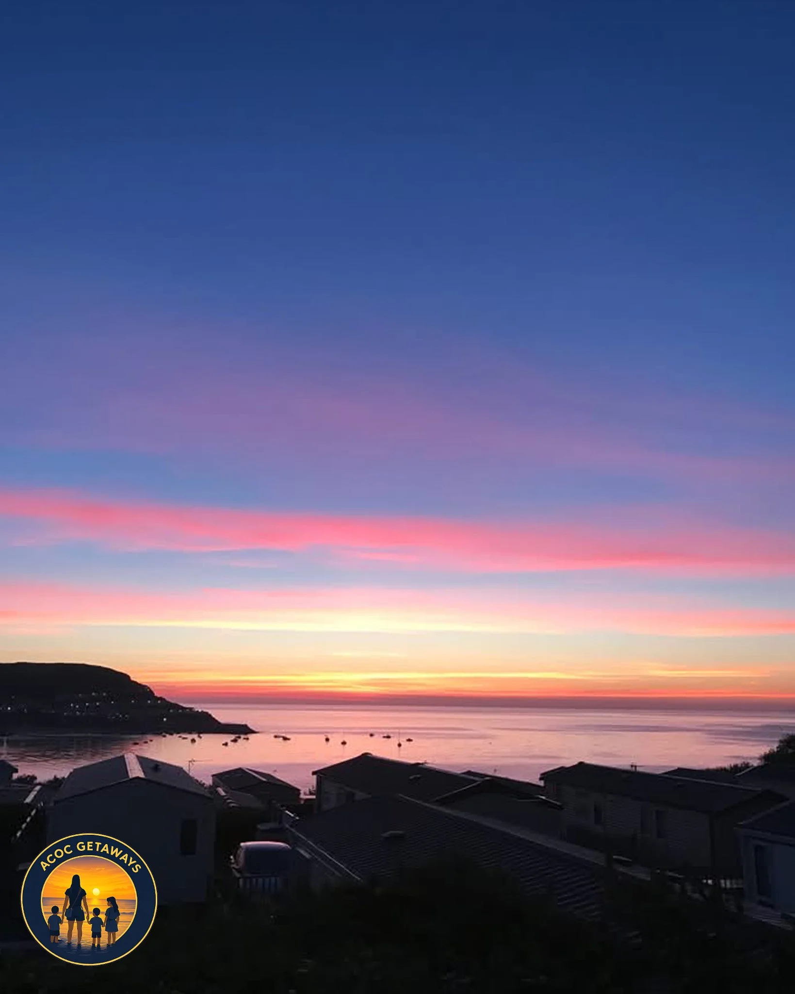 Colorful sunset over a calm body of water, with silhouettes of houses and a hill in the foreground, and boats on the water.