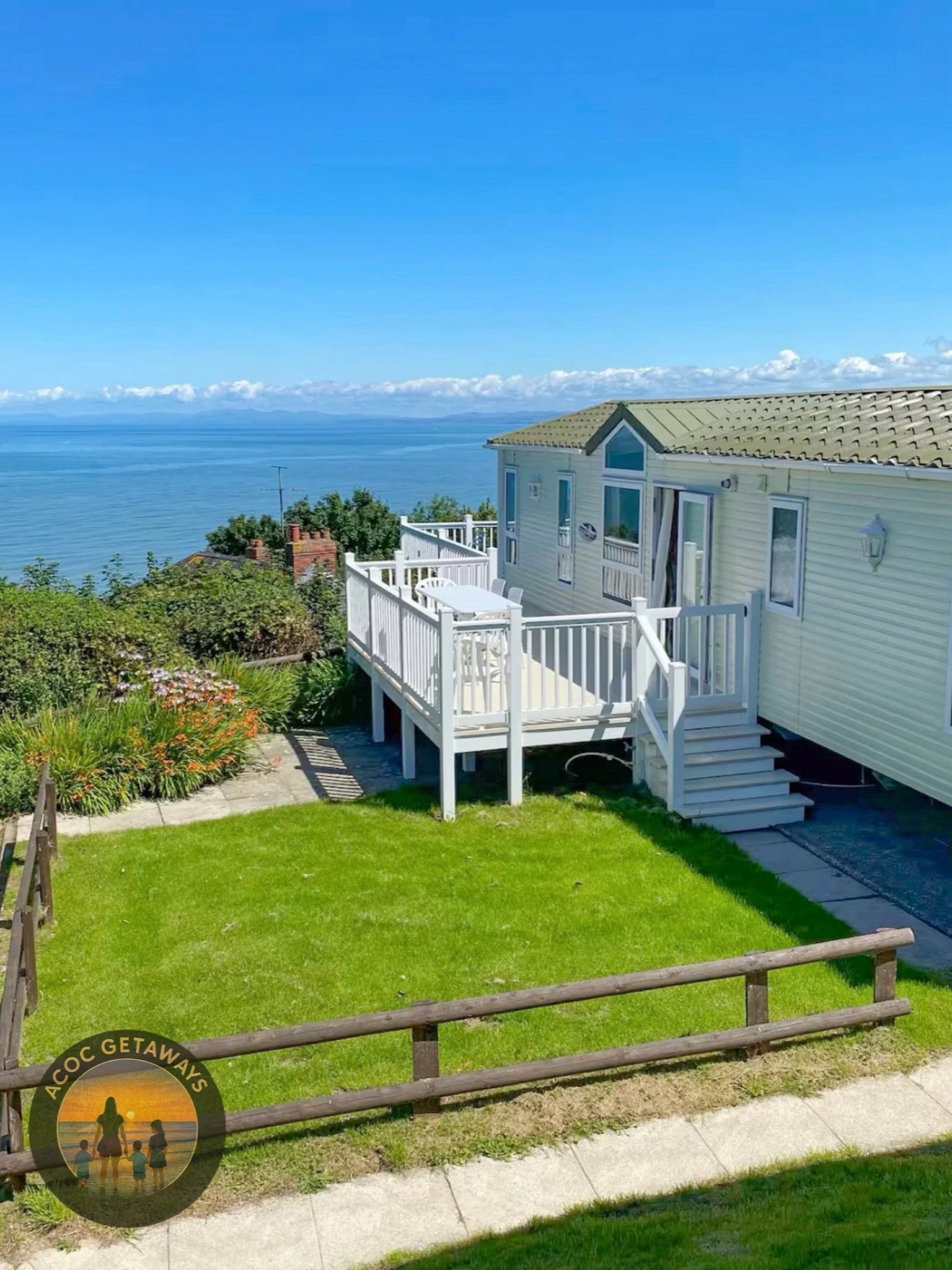 A seaside house with a white deck, yellow siding, and a view of the ocean with blue sky and clouds, green lawn, flower bed, and a wooden fence in the foreground.