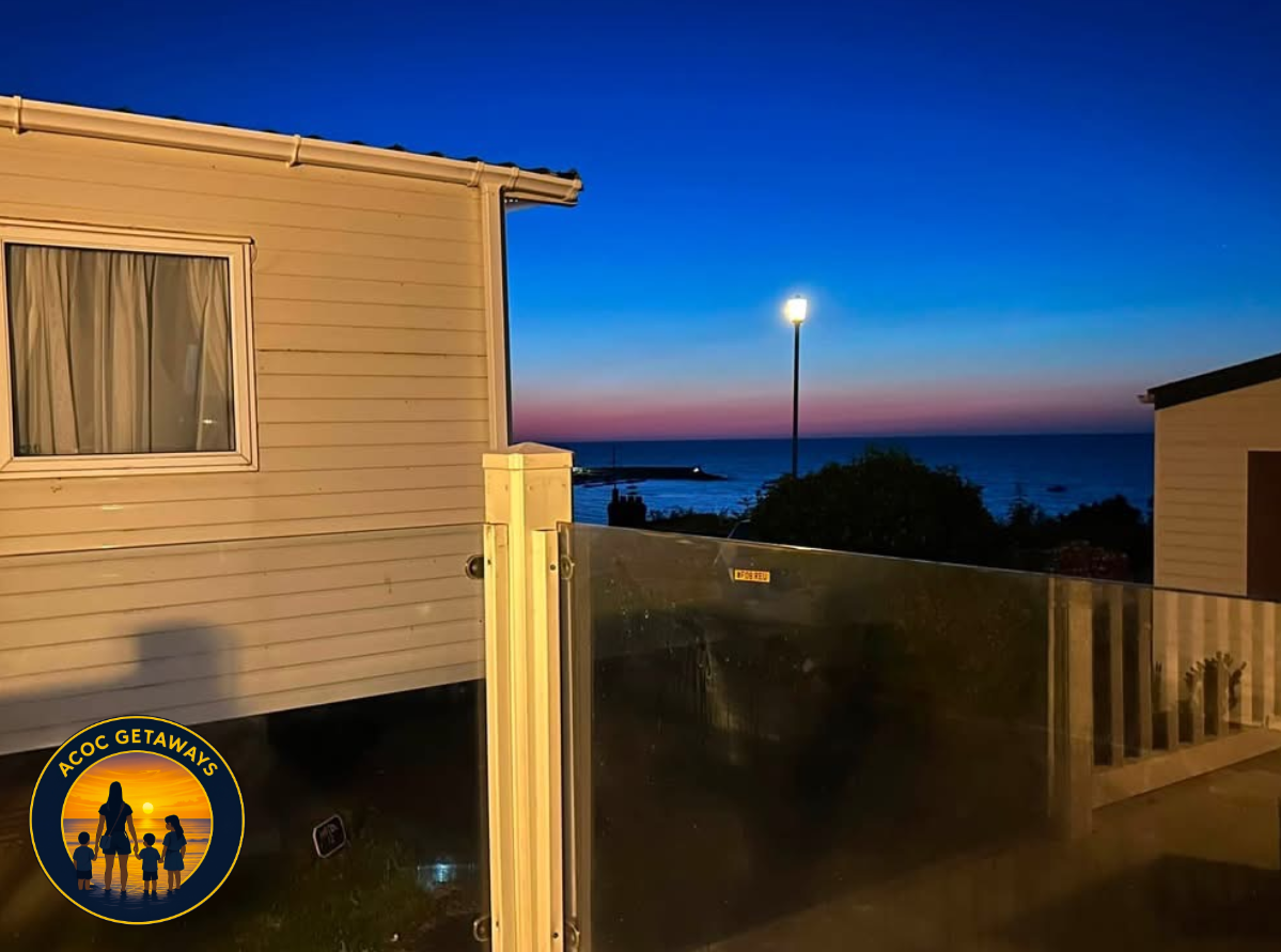 View from a balcony at dusk, showing a house with beige siding and a window with closed curtains, a streetlamp, a fence, and the ocean with a colorful sky in the background.