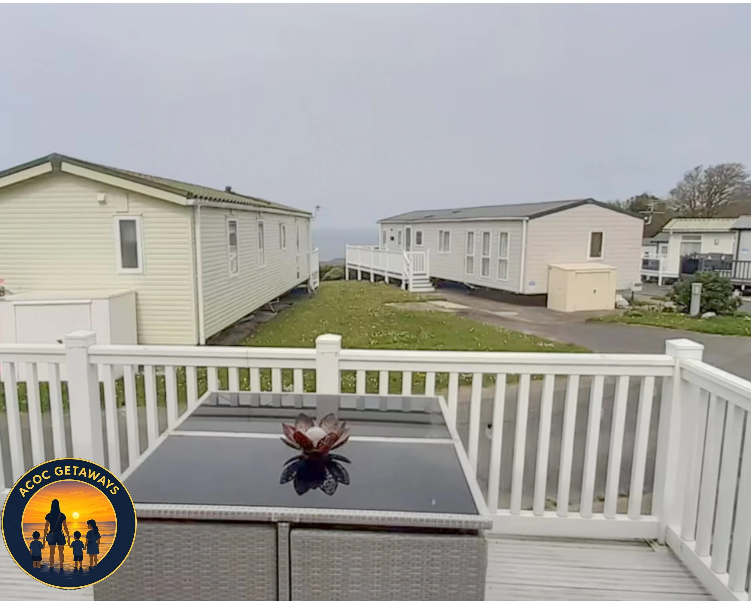 A balcony with a table and a decorative flower, overlooking mobile homes on a cloudy day with greenery and trees in the background.