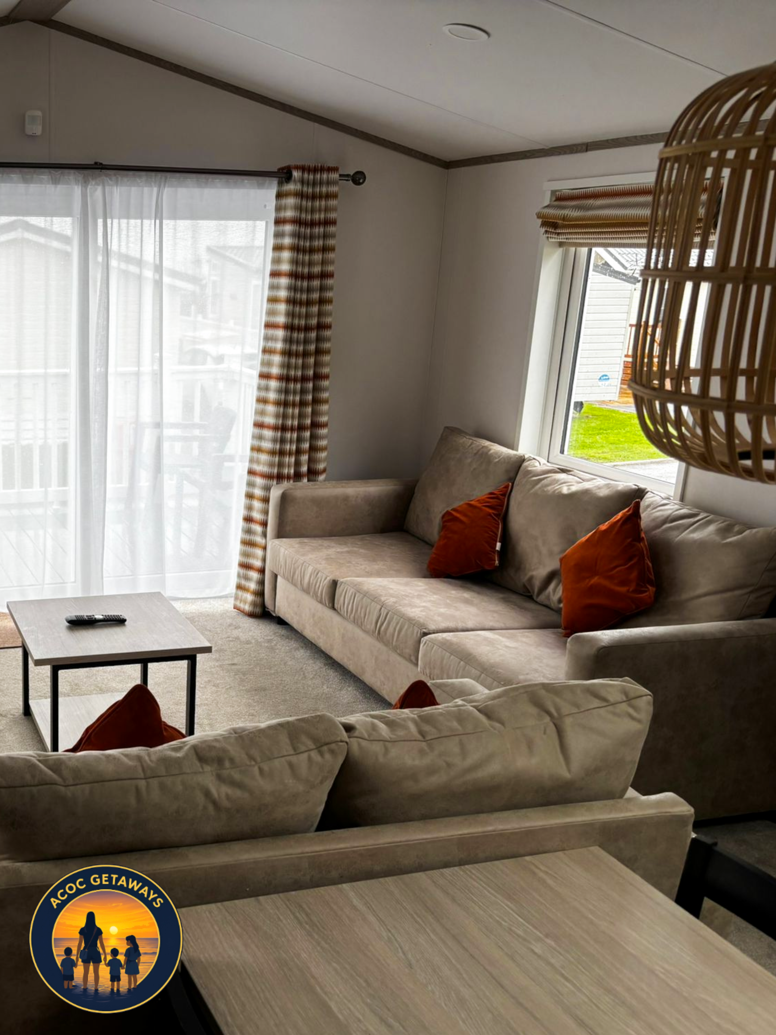 Living room with beige sofas, orange pillows, a small rectangular coffee table, large windows with white and patterned curtains, a hanging light fixture, and a view of a backyard with grass.