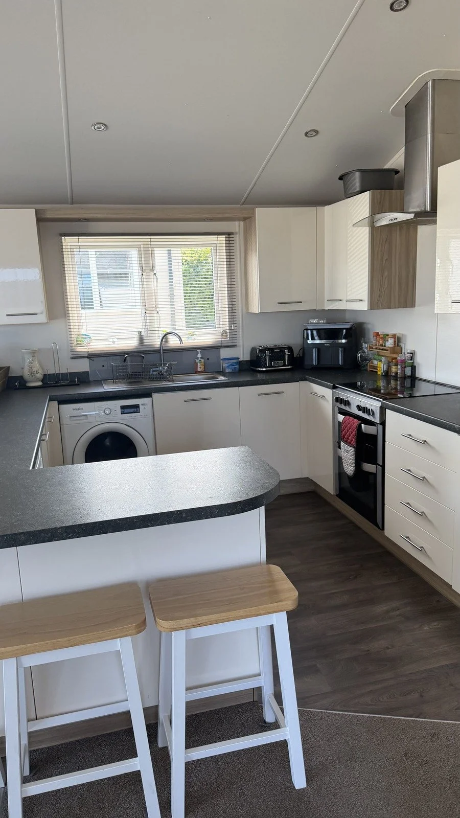 Kitchen with white cabinets, black countertops, stainless steel appliances, a window above the sink, and two wooden bar stools at a breakfast bar.