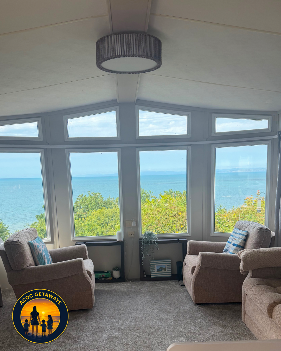 A cozy living room with large bay windows overlooking the ocean and green foliage, furnished with beige armchairs with striped and patterned cushions, a dark floor-based shelf with decor, and a ceiling light fixture.
