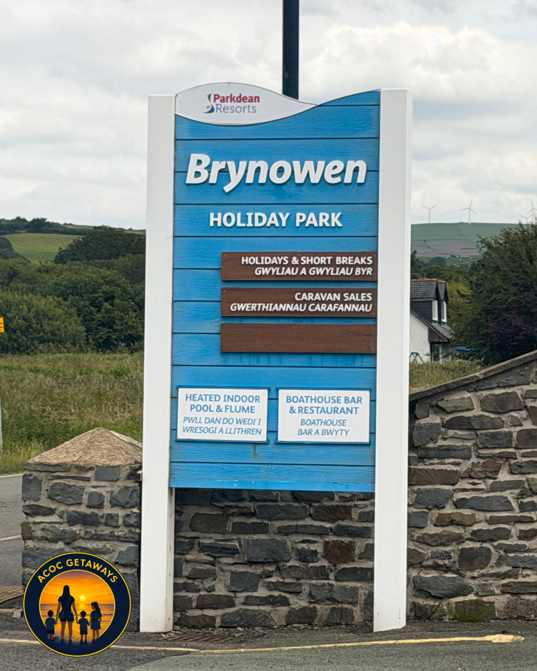 Signboard for Brynowen Holiday Park with information about indoor pool, bar, restaurant, and caravan sales, located outdoors with greenery and wind turbines in the background.