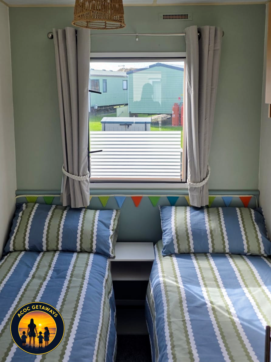 A cozy bedroom with two twin beds with blue, green, and white striped bedding, a window with white curtains and a view of neighboring houses, a small white bedside table between the beds, and a wicker lampshade hanging from the ceiling.