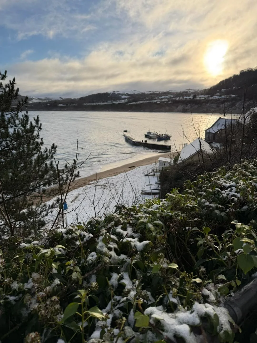 Snow-covered beach with boats in the water, trees and bushes in the foreground, hills in the background, and a cloudy sky with the sun partially visible.