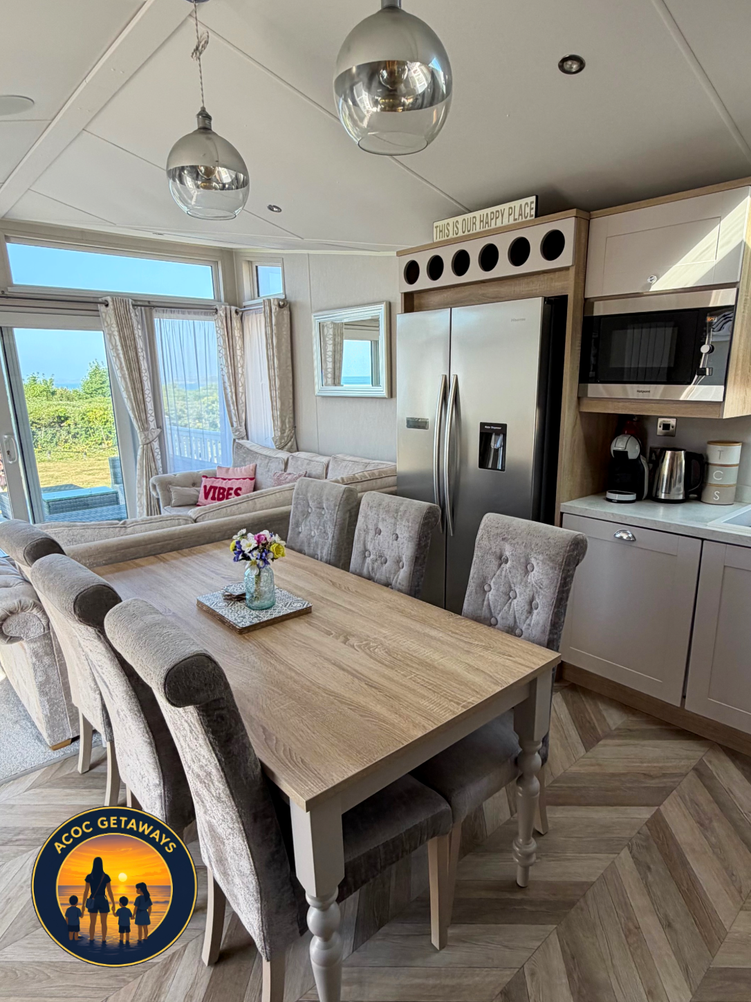Open-concept living and kitchen area with dining table, upholstered chairs, a refrigerator, microwave, and a view through sliding glass doors to outdoor greenery.