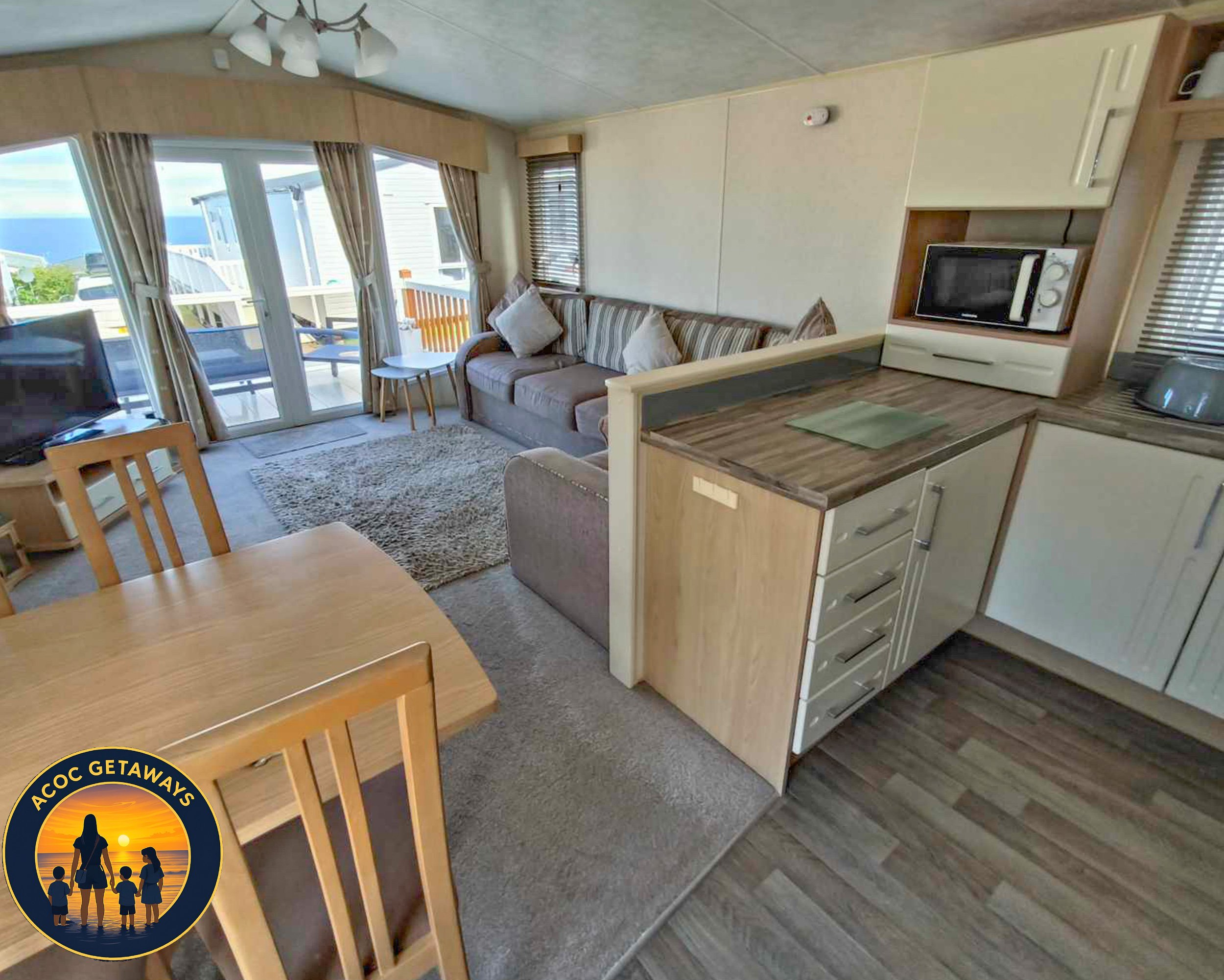 Living area with sliding glass door leading to balcony, striped sofa with pillows, wooden dining table and chairs, small rug, TV, microwave, and kitchen counter with cabinets in a cozy apartment.