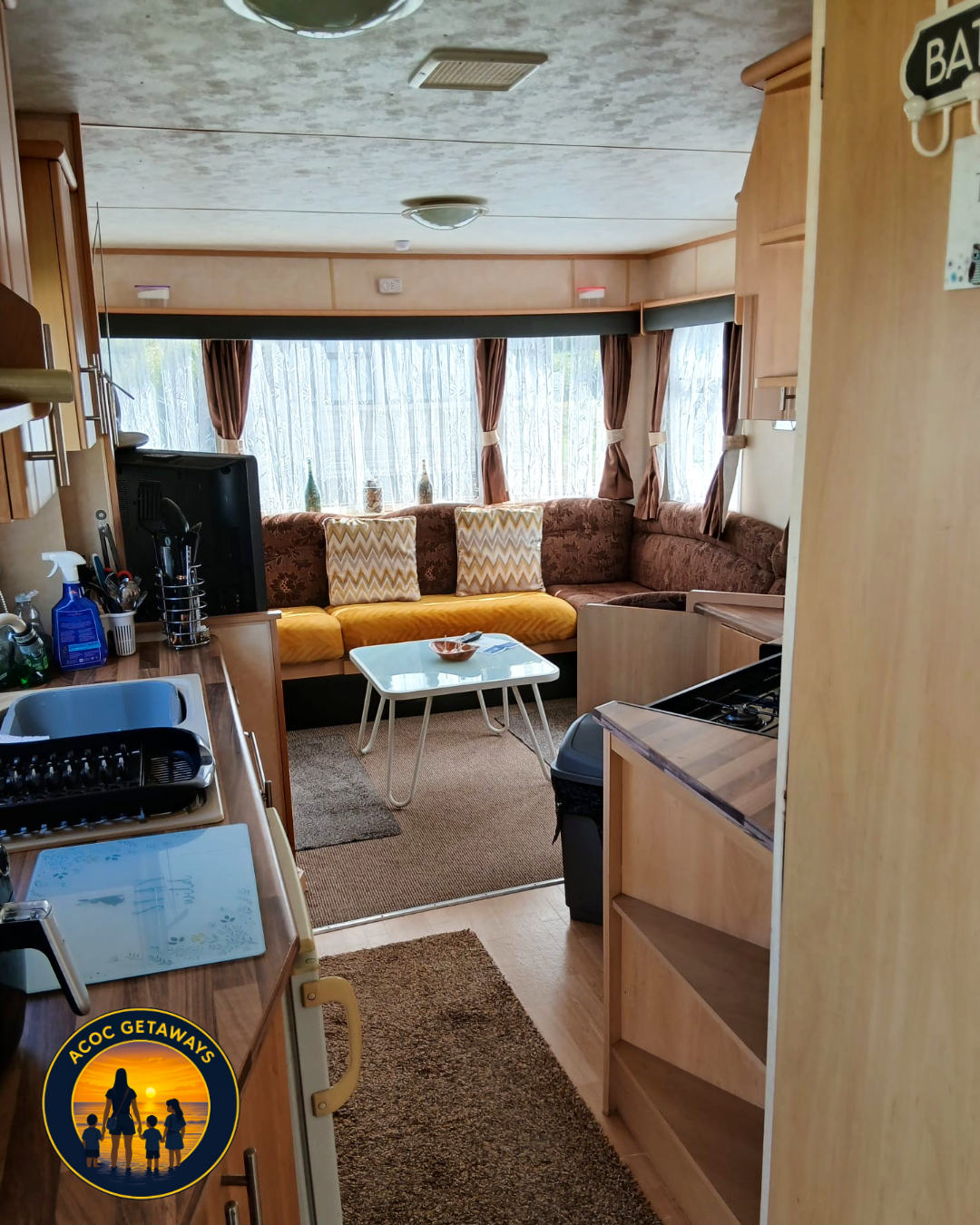 Interior view of a cozy living space with a kitchen area on the left and a seating area with a sofa and large windows with curtains in the background.