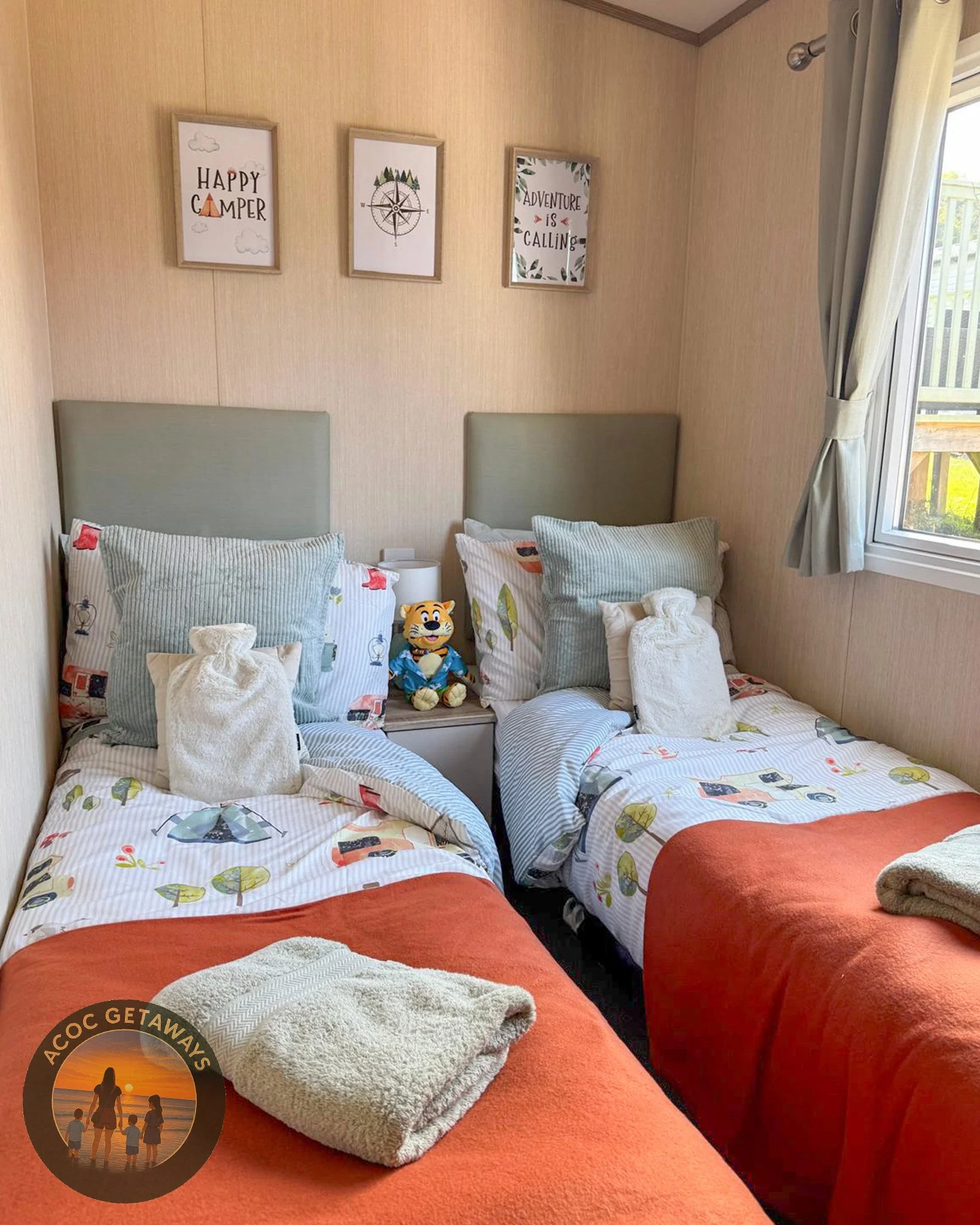 A cozy bedroom with two single beds featuring colorful camping-themed bedding, each with multiple pillows and a towel on top. A plush tiger toy is placed on the nightstand between the beds. The wall above the beds has three framed quotes with nature 