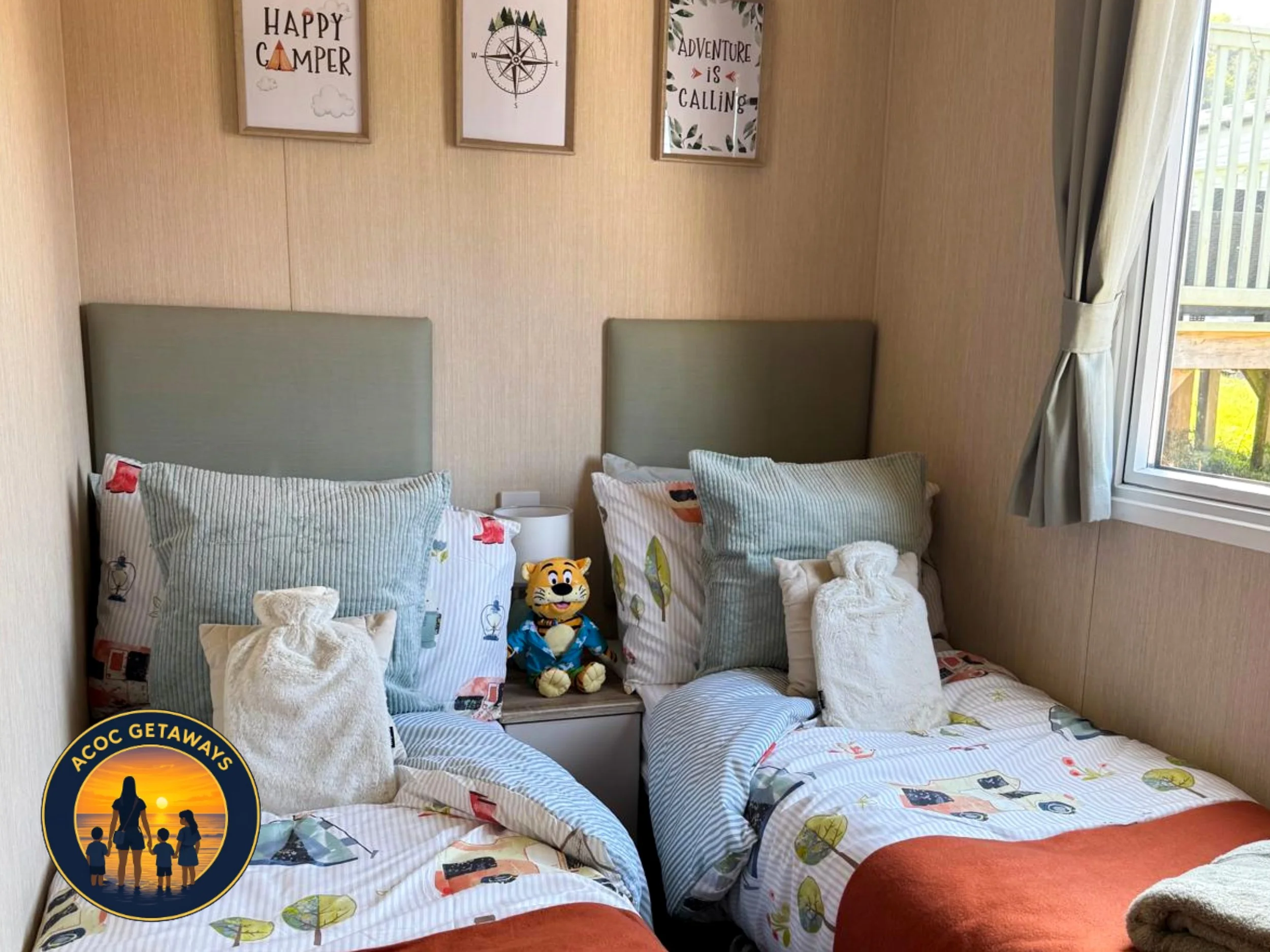 A cozy bedroom with a bed covered in sailboat and travel-themed bedding. Multiple pillows are arranged on the bed, and plush toys, including a tiger character, are placed at the head of the bed. A window with curtains allows natural light into the ro