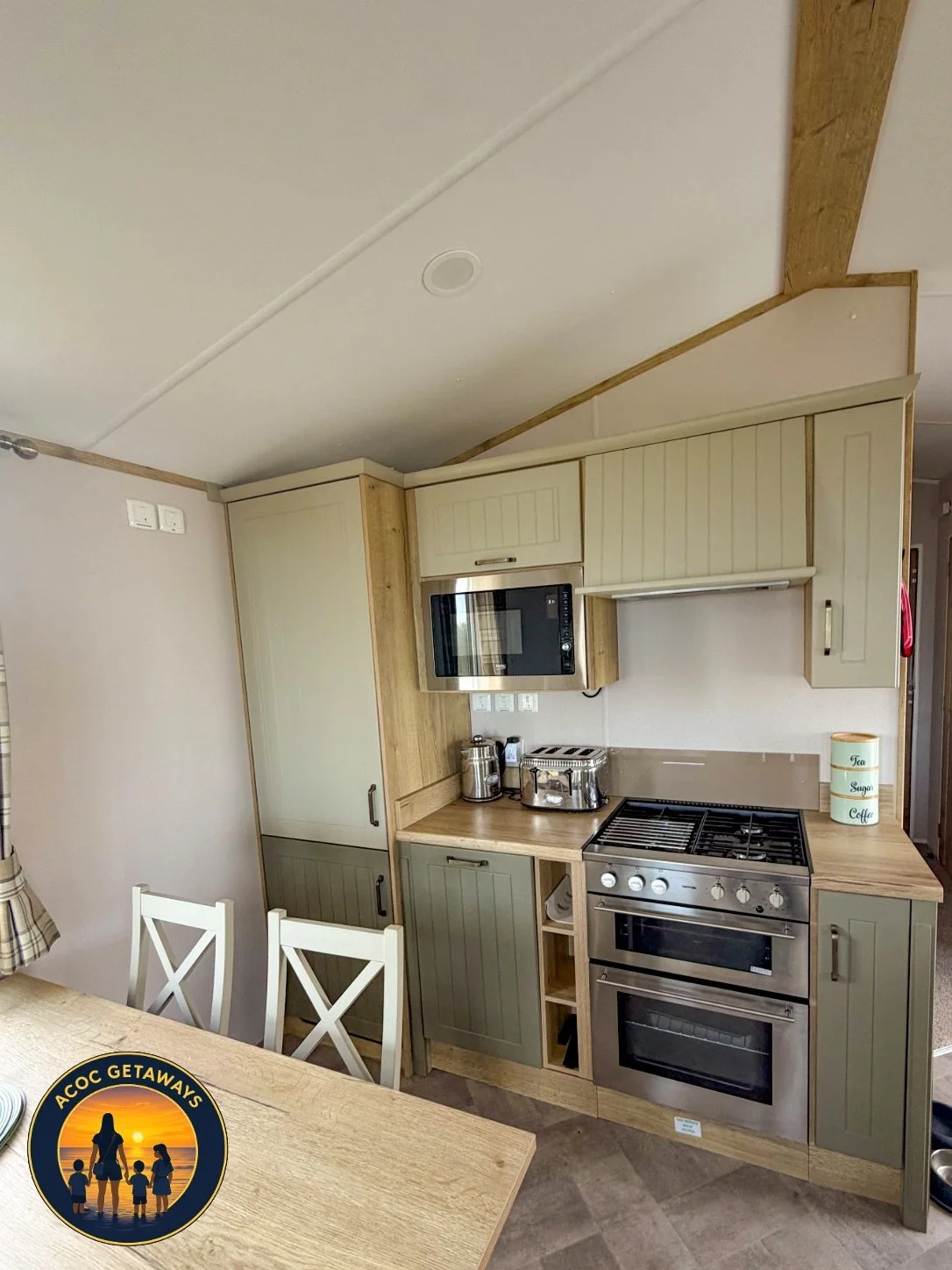 Kitchen with beige cabinets, stainless steel stove and oven, microwave, and small appliances on the counter. Part of a dining table with white chairs and a logo on the image that reads "ACOC Getaways" with a silhouette of a family at the beach.