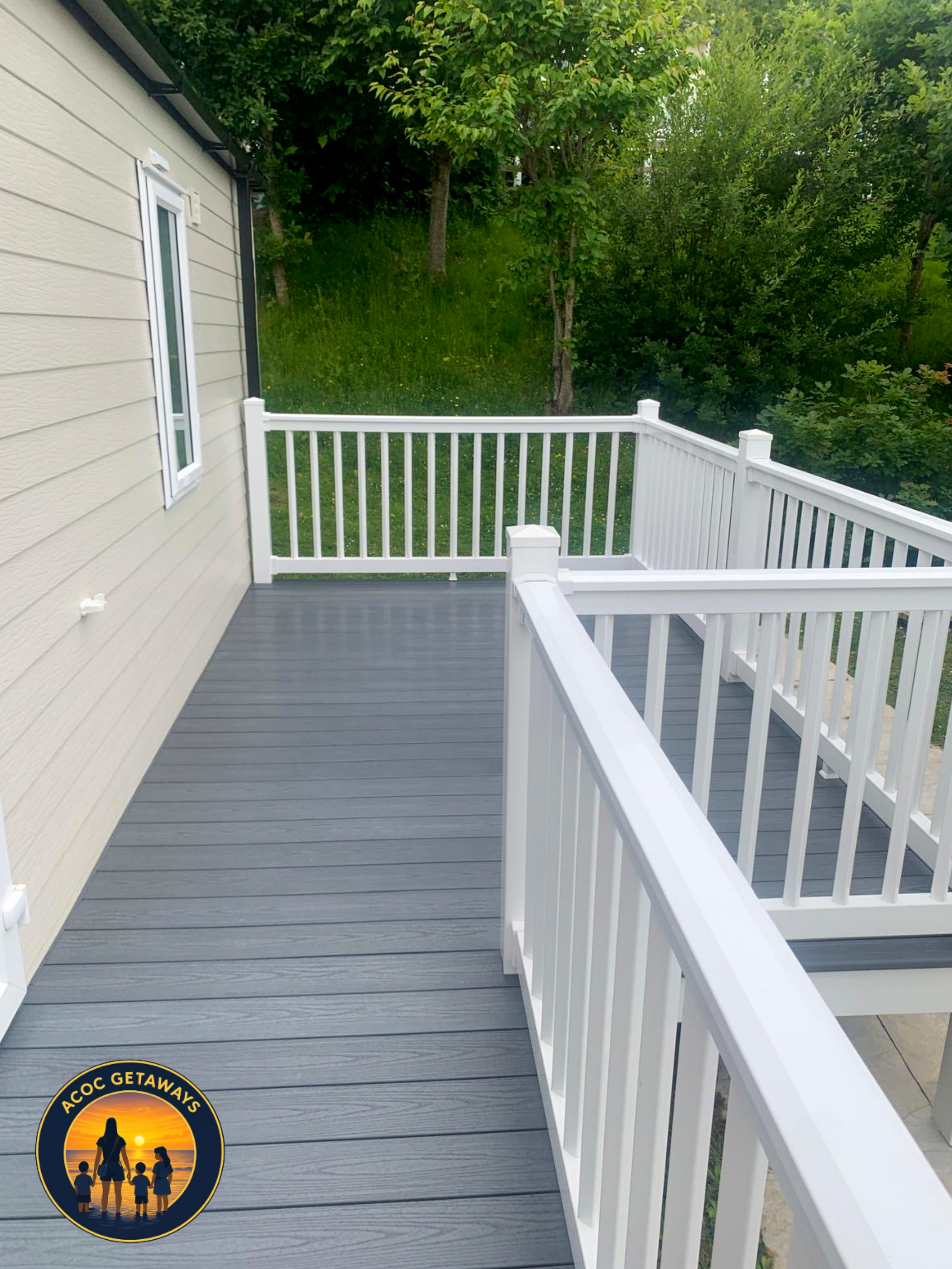 A view from a balcony with grey wooden decking, surrounded by white railing. To the left, part of a beige house wall with a small window. In the background, a grassy backyard with several trees and green foliage.