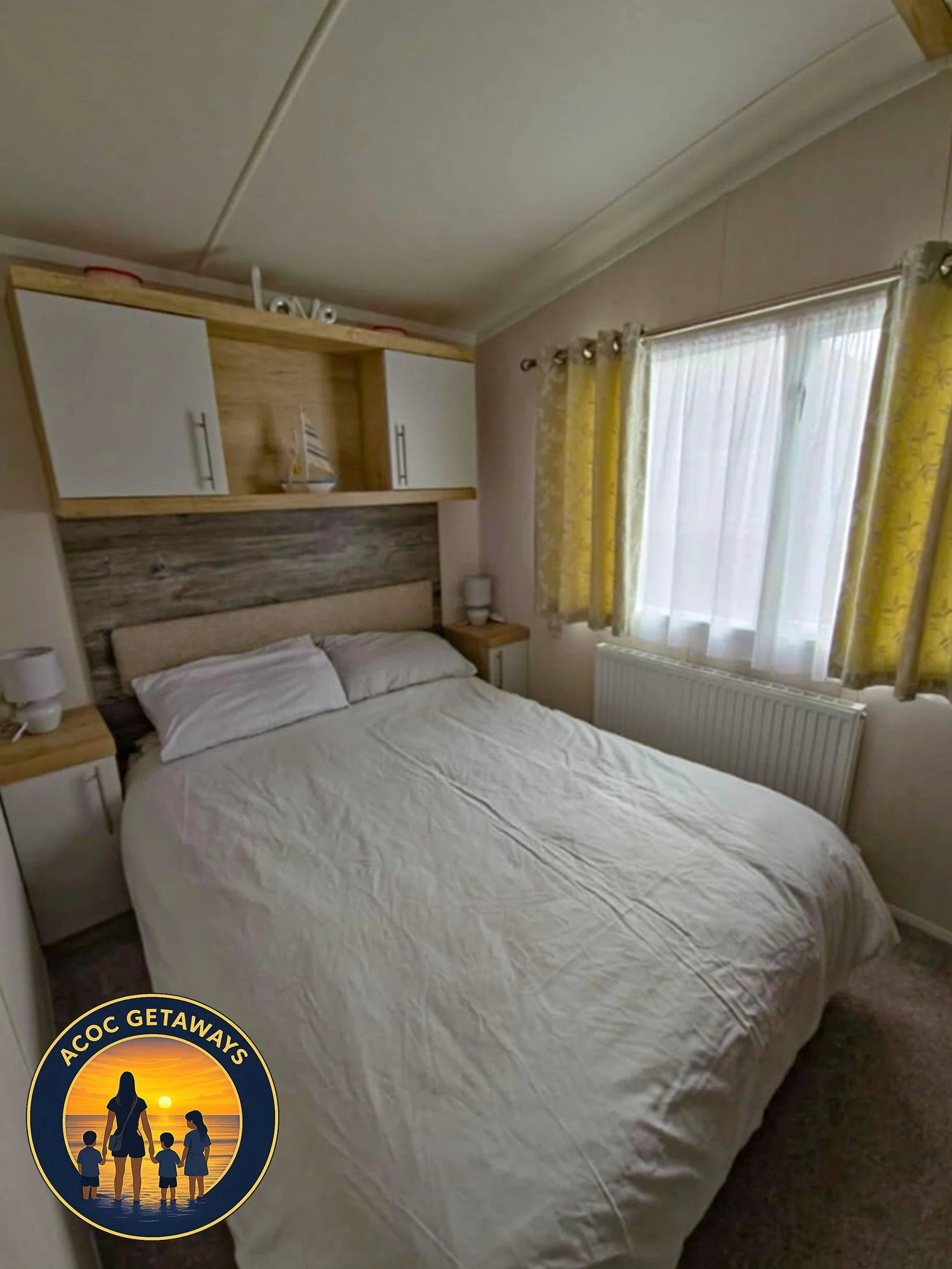A cozy bedroom with a white bed, wooden headboard, and matching nightstands. There is a window with yellow curtains and white sheer curtains, allowing natural light to brighten the room. An ACoc Getaways logo is present at the bottom left corner of the image.